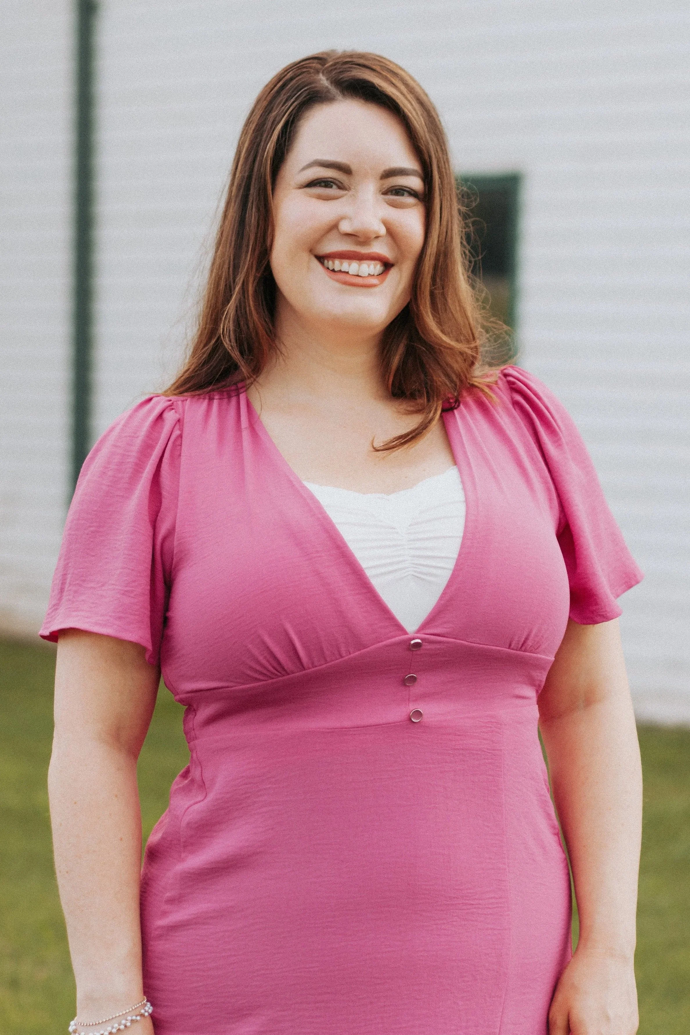 A smiling woman with shoulder-length brown hair wearing a pink dress and standing outdoors in front of a white building.