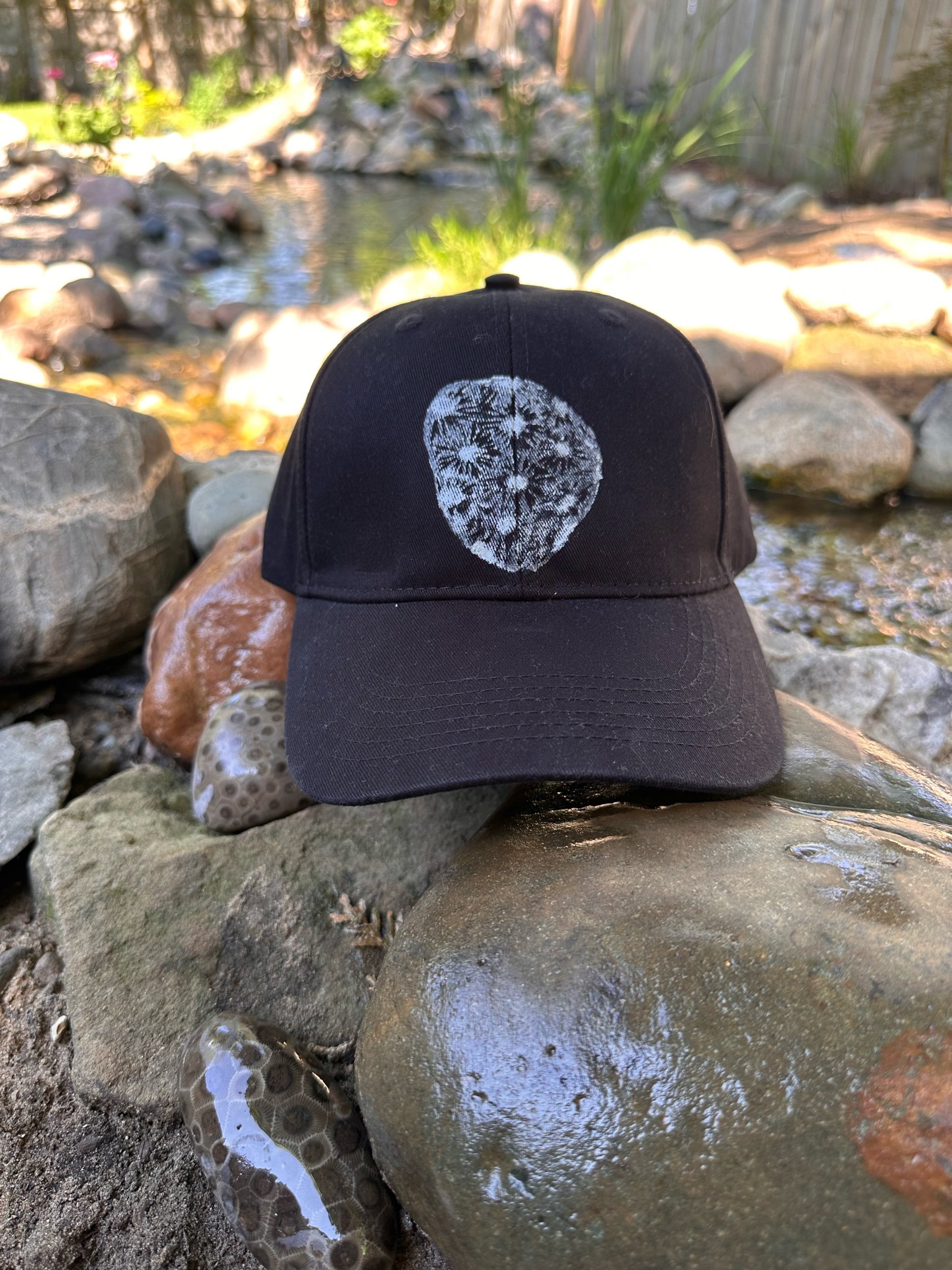 Black hat with a white pearl hand-printed Petoskey stone design, resting on a rock beside real Petoskey stones near a backyard koi pond.