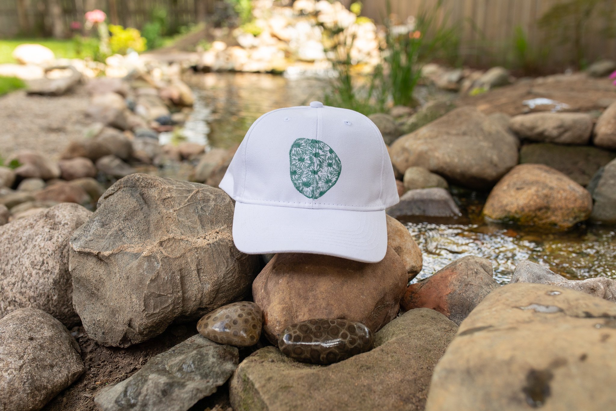 White hat with a hand-printed Petoskey stone design in teal ink, displayed outdoors near natural stones and greenery.