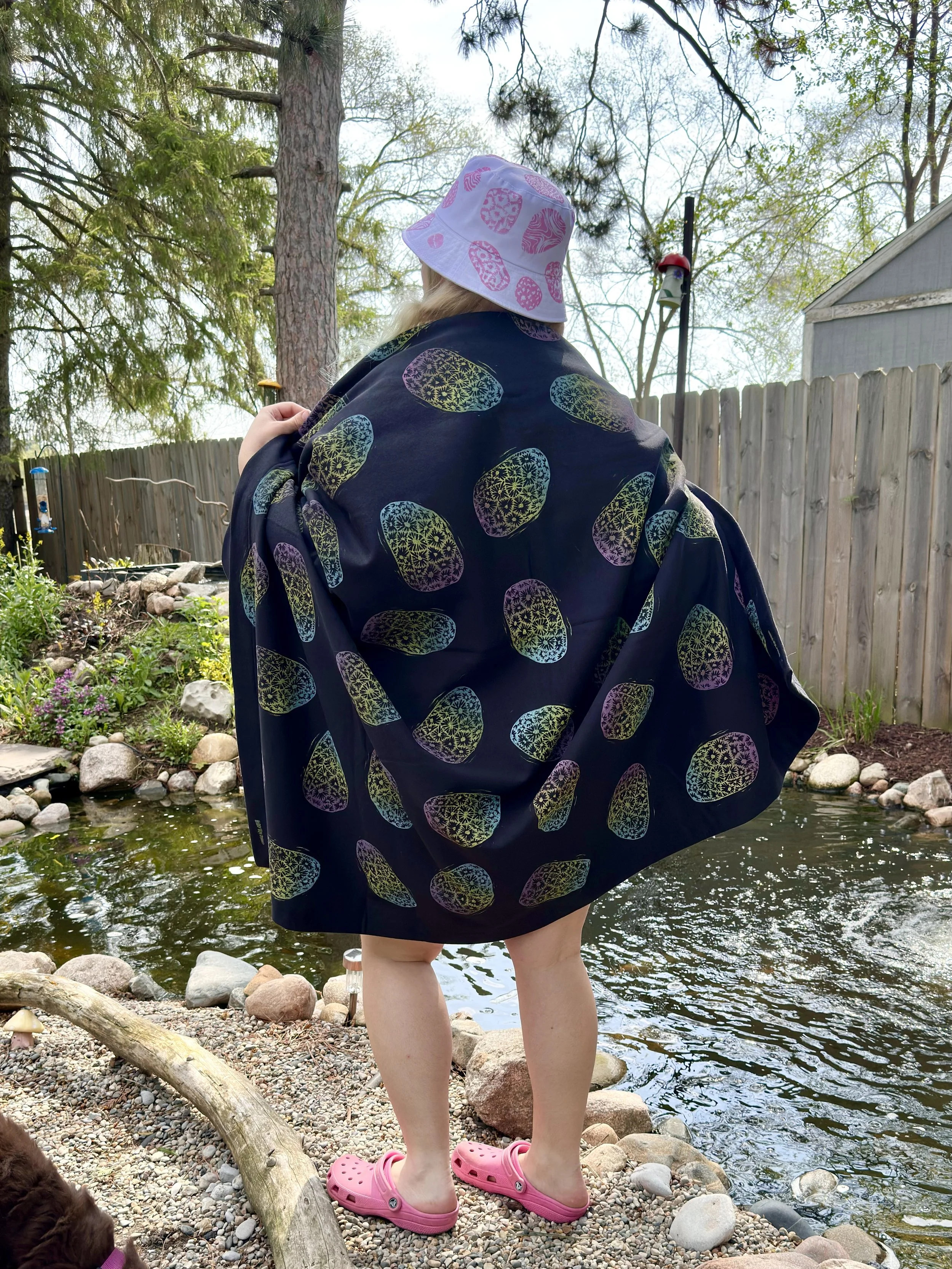 A person wearing a white sun hat with pink patterns and a dark blue blanket with colorful petoskey stone designs, standing on rocks by a pond. The limbs of a small dog in pink shoes are partially visible in the lower left corner.