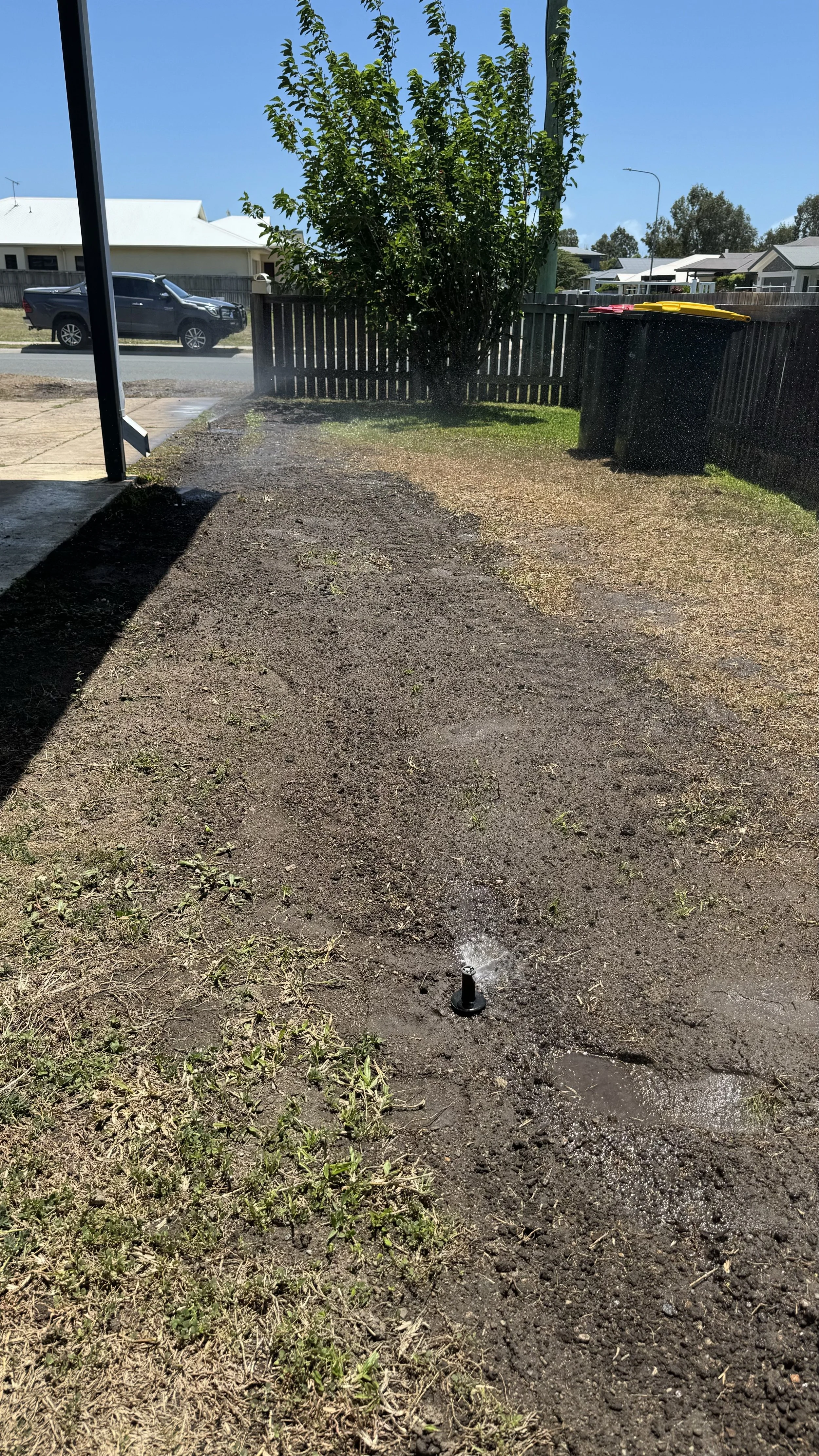 A backyard with a patch of dirt and grass, a tree, trash bins, and a water sprinkler spraying water on the ground.
