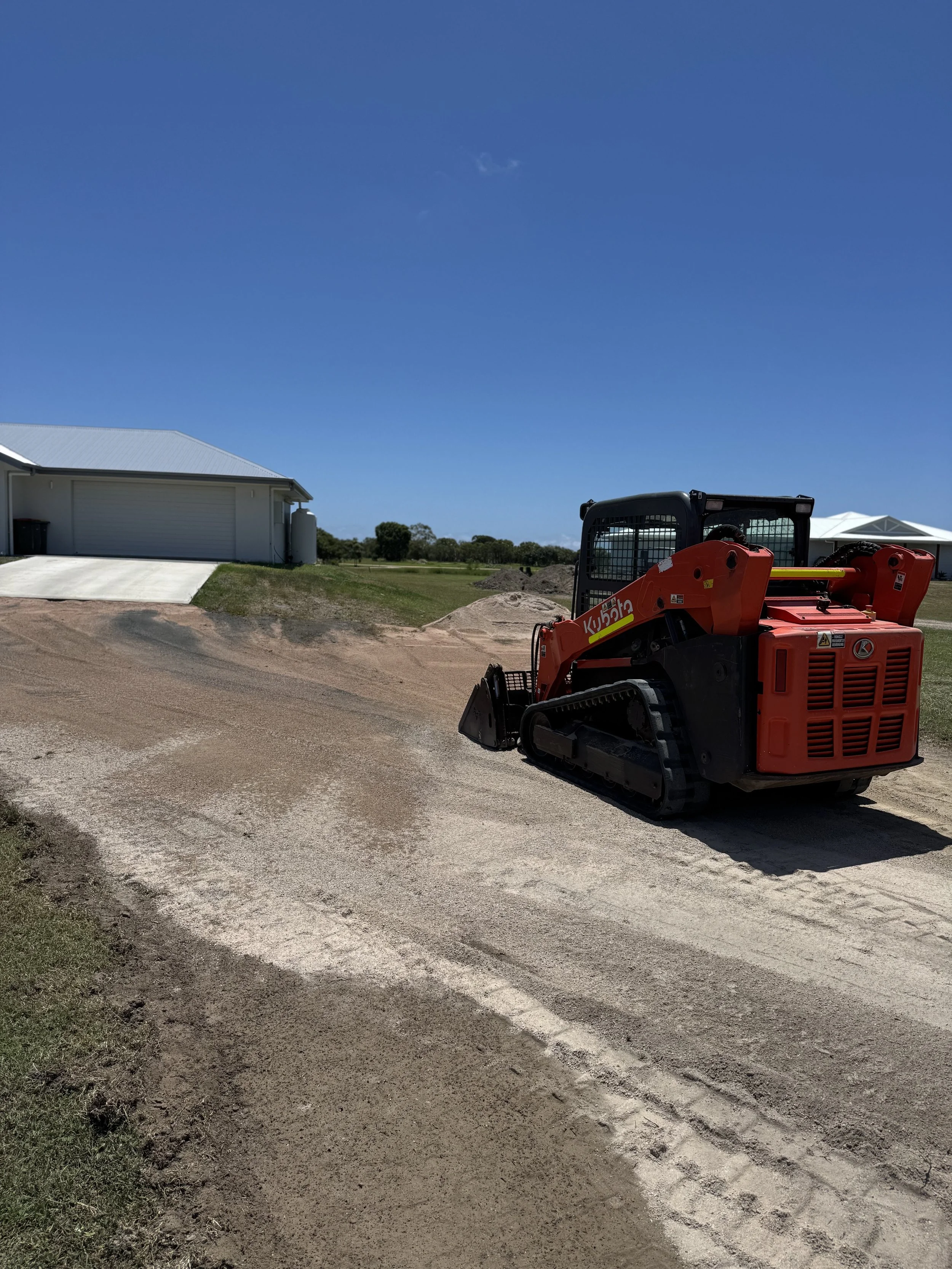 A small red compact excavator working on a sandy driveway in front of a house with a white roof and a metal garage door, under a clear blue sky.