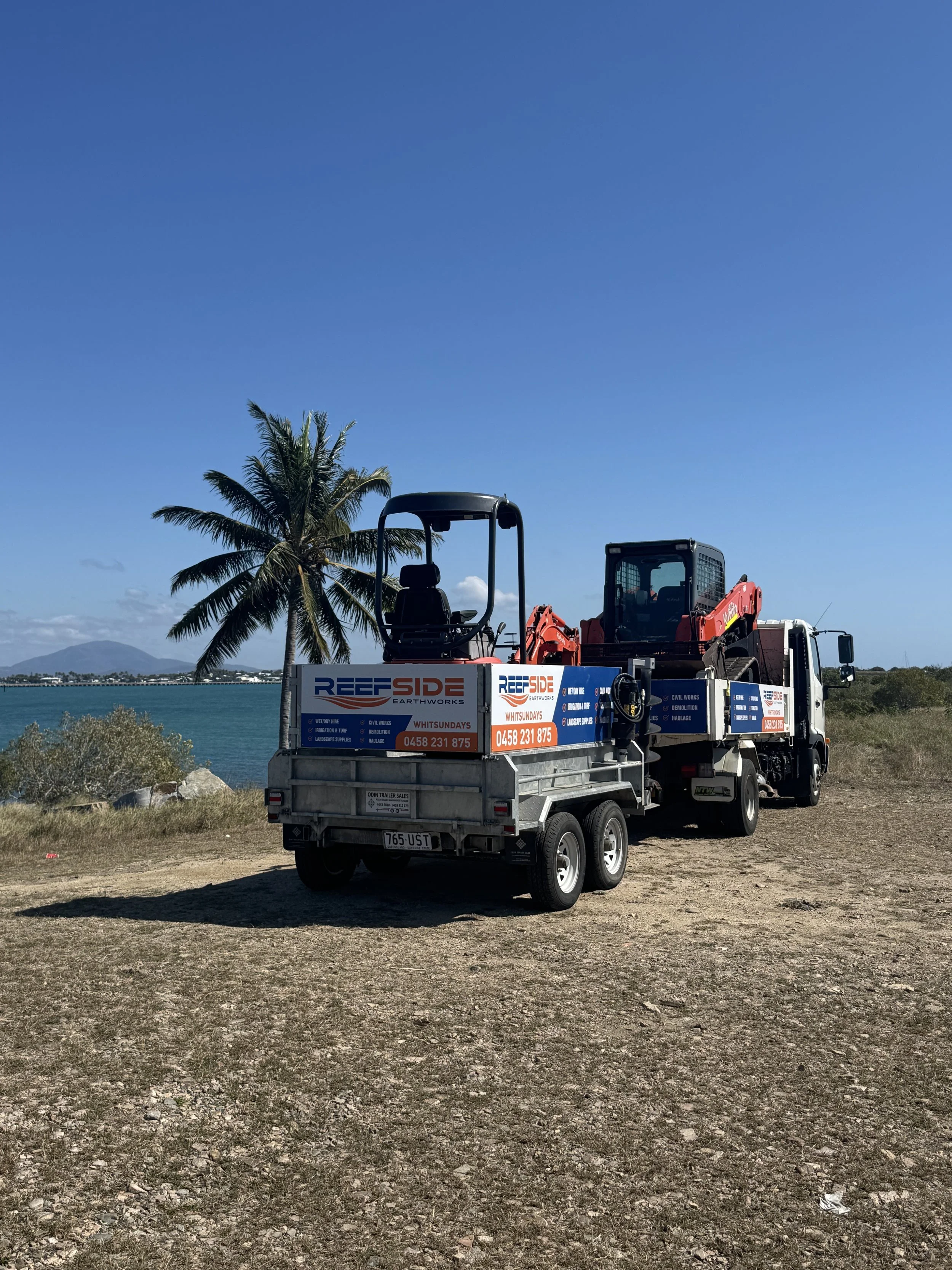 A truck with a smaller trailer attached, parked on a rocky dirt area near a body of water with palm trees and distant mountains in the background. The truck and trailer are branded with 'Reefside Earthworks' and contact information for Whitsundays.