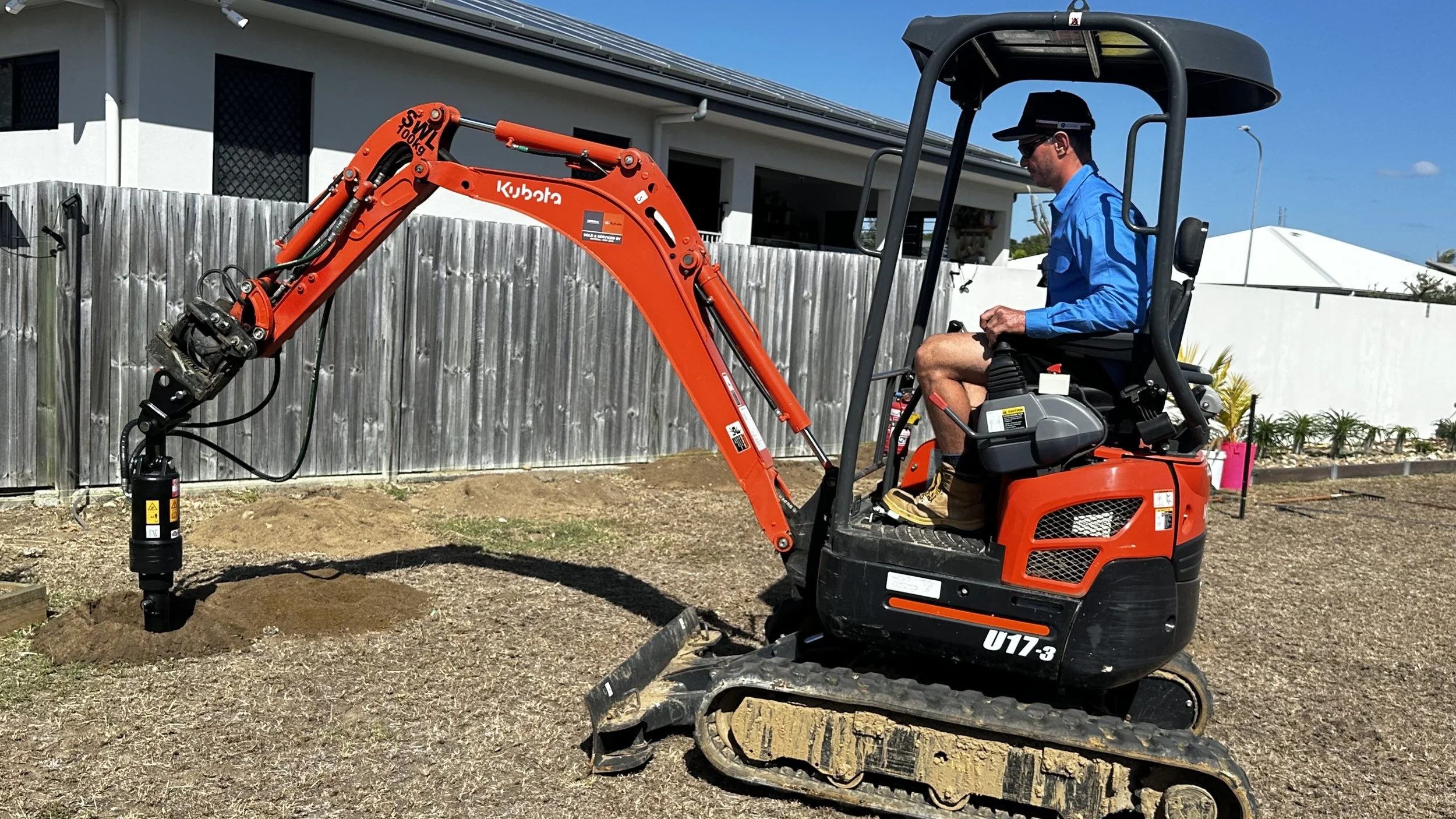 A man operating a small orange Kubota mini excavator digging into the ground in a backyard, with a wooden fence and house in the background, under a blue sky.