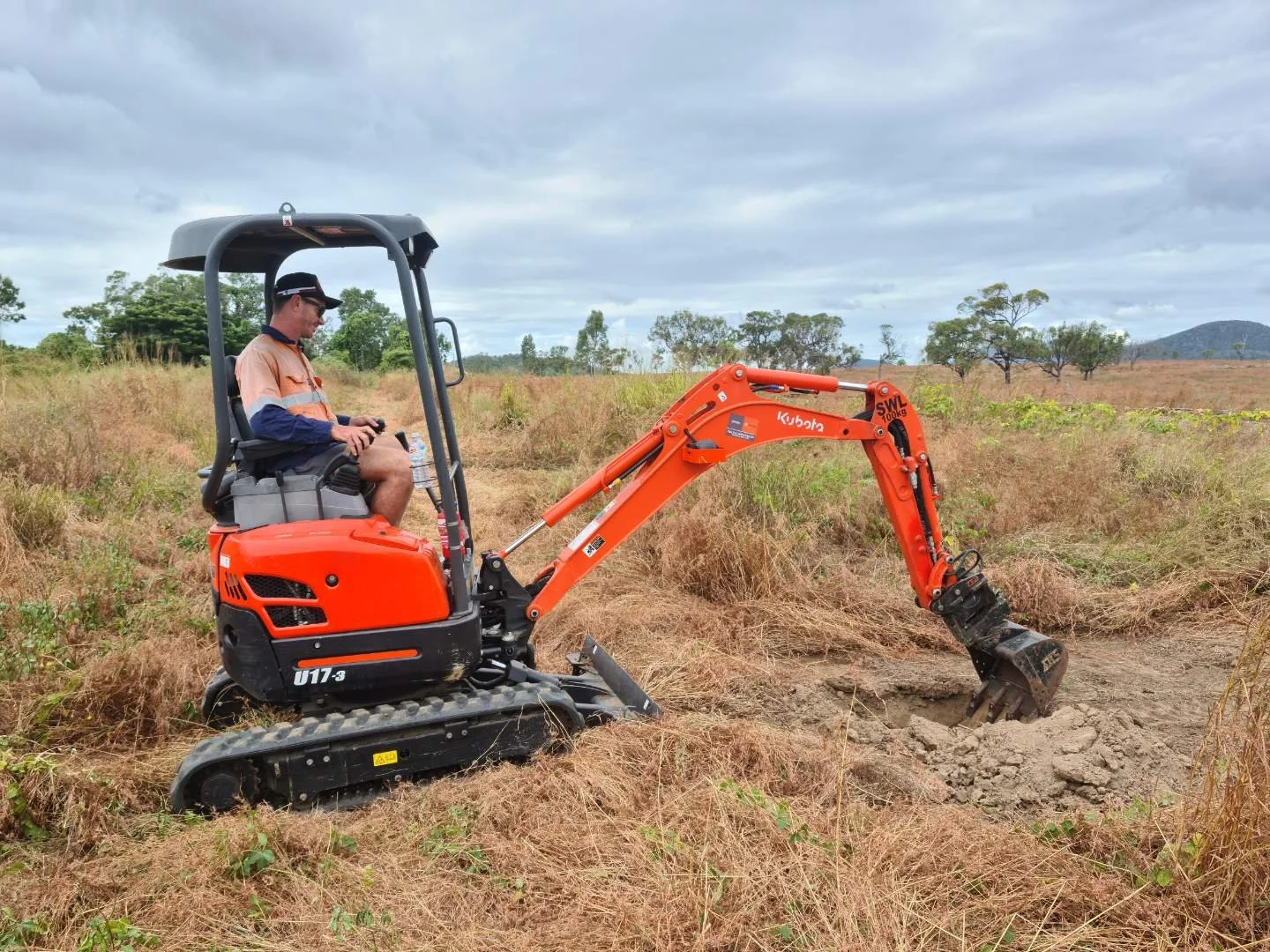 Man operating an orange Kubota mini excavator on a grassy field with cloudy sky and trees in the background.