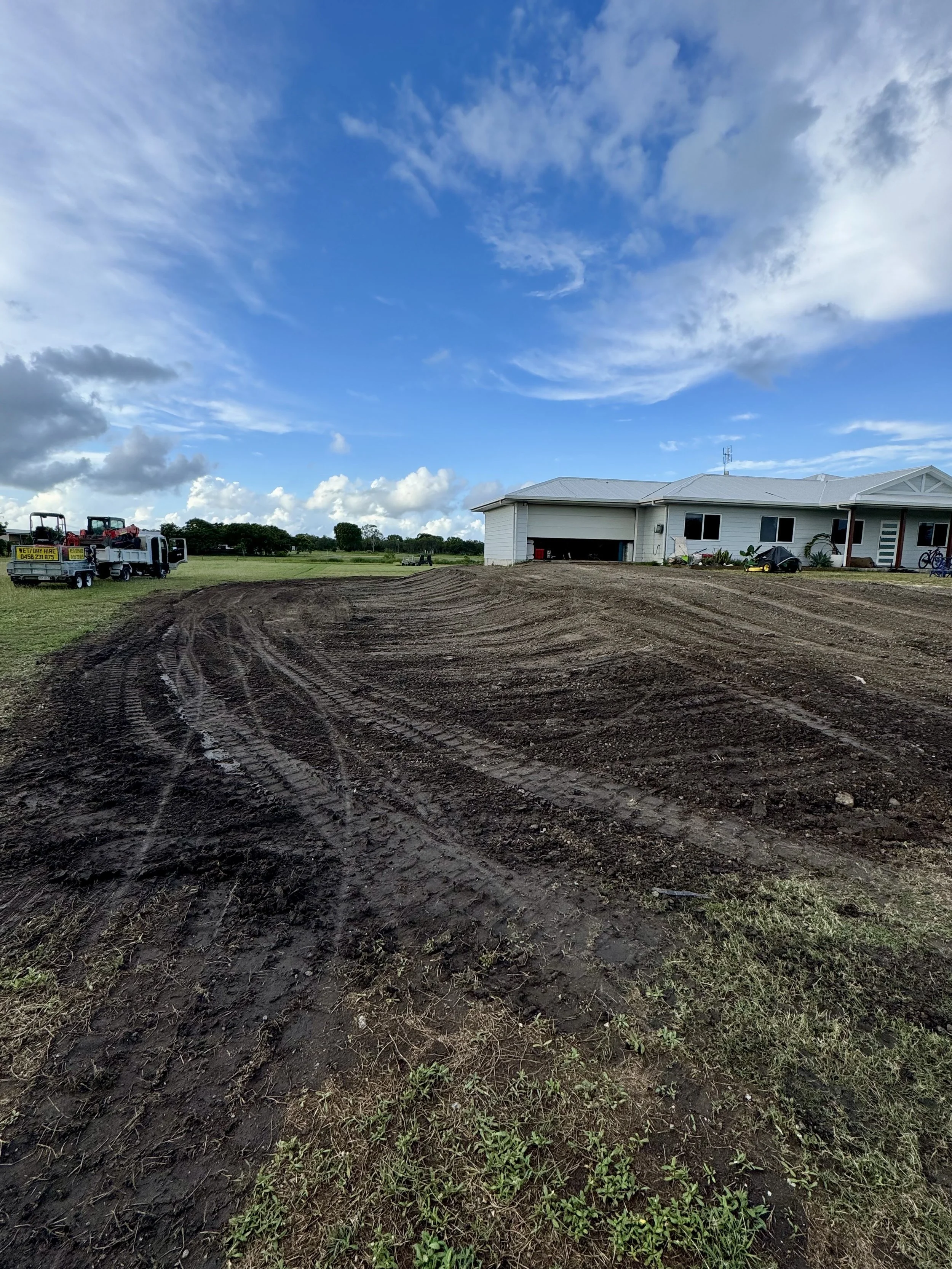 A house under construction on a dirt lot with construction vehicles parked nearby, and a blue sky with clouds overhead.