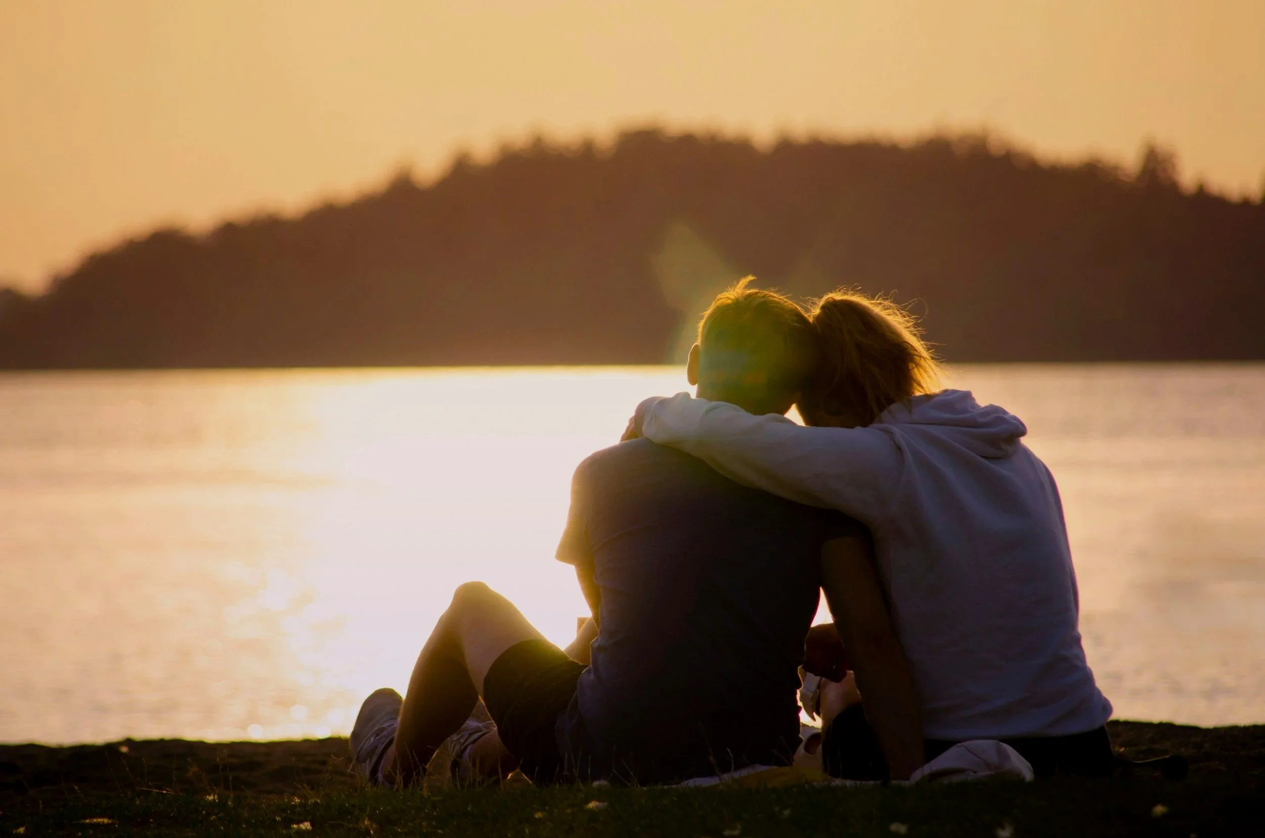 A couple sitting close together on the beach at sunset, with their heads touching, overlooking a body of water and a silhouetted island or hill in the background.