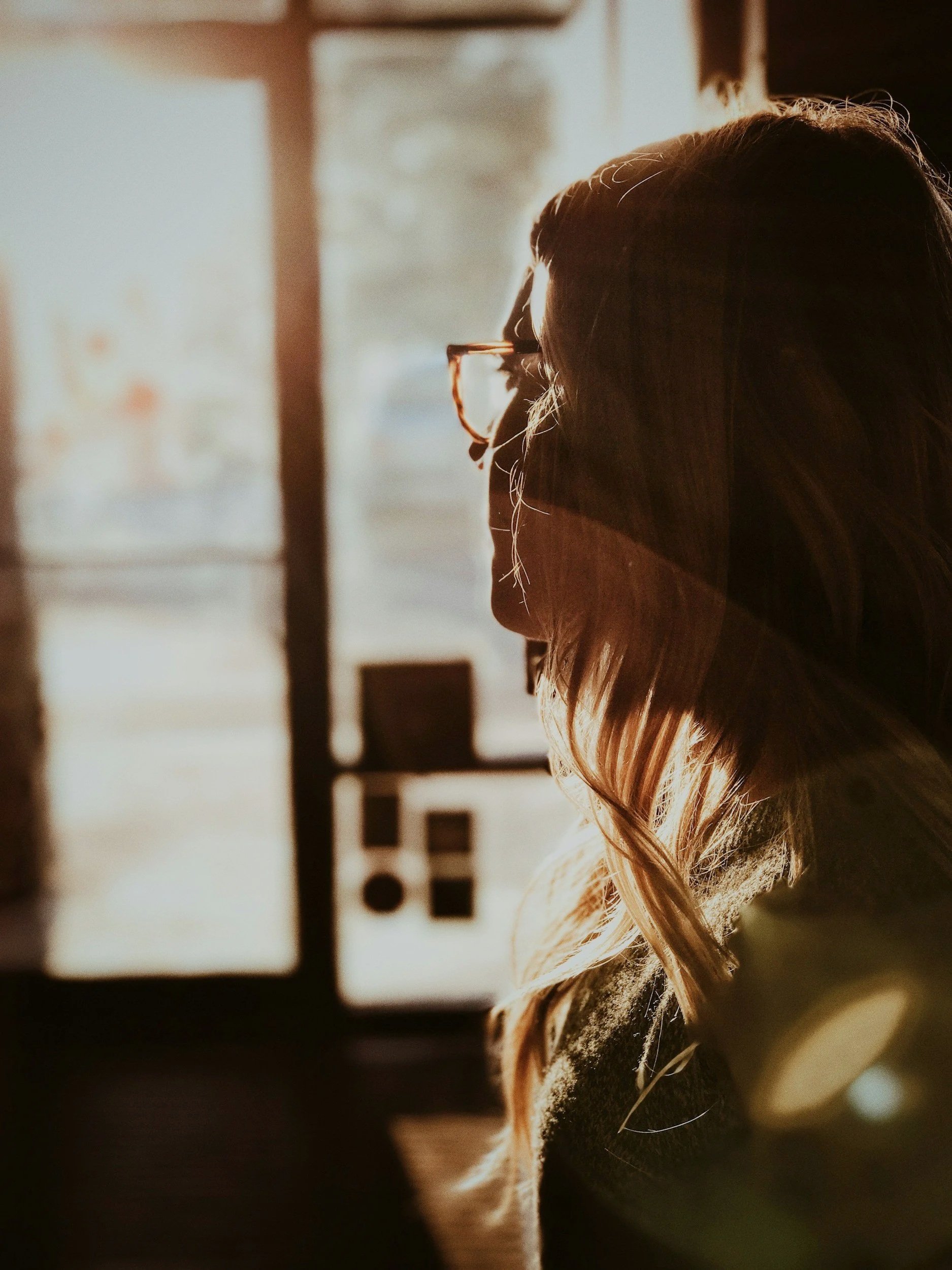 A woman with glasses is sitting indoors, illuminated by sunlight coming through a window, creating a warm glow on her profile.