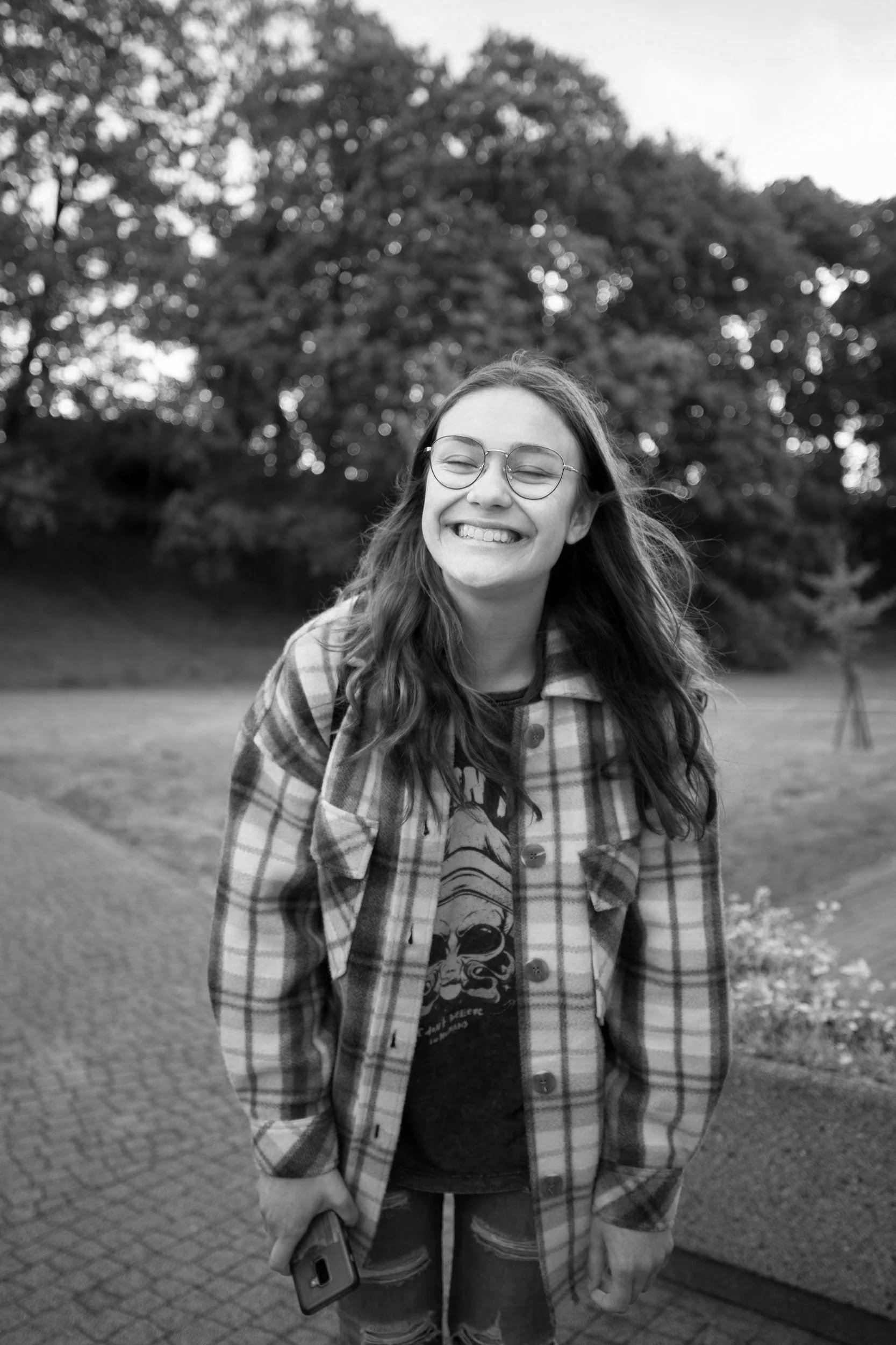 A young woman with long hair, glasses, and a big smile stands outdoors in a park, holding a phone in her right hand, wearing a plaid shirt over a graphic t-shirt, with trees and grass in the background.