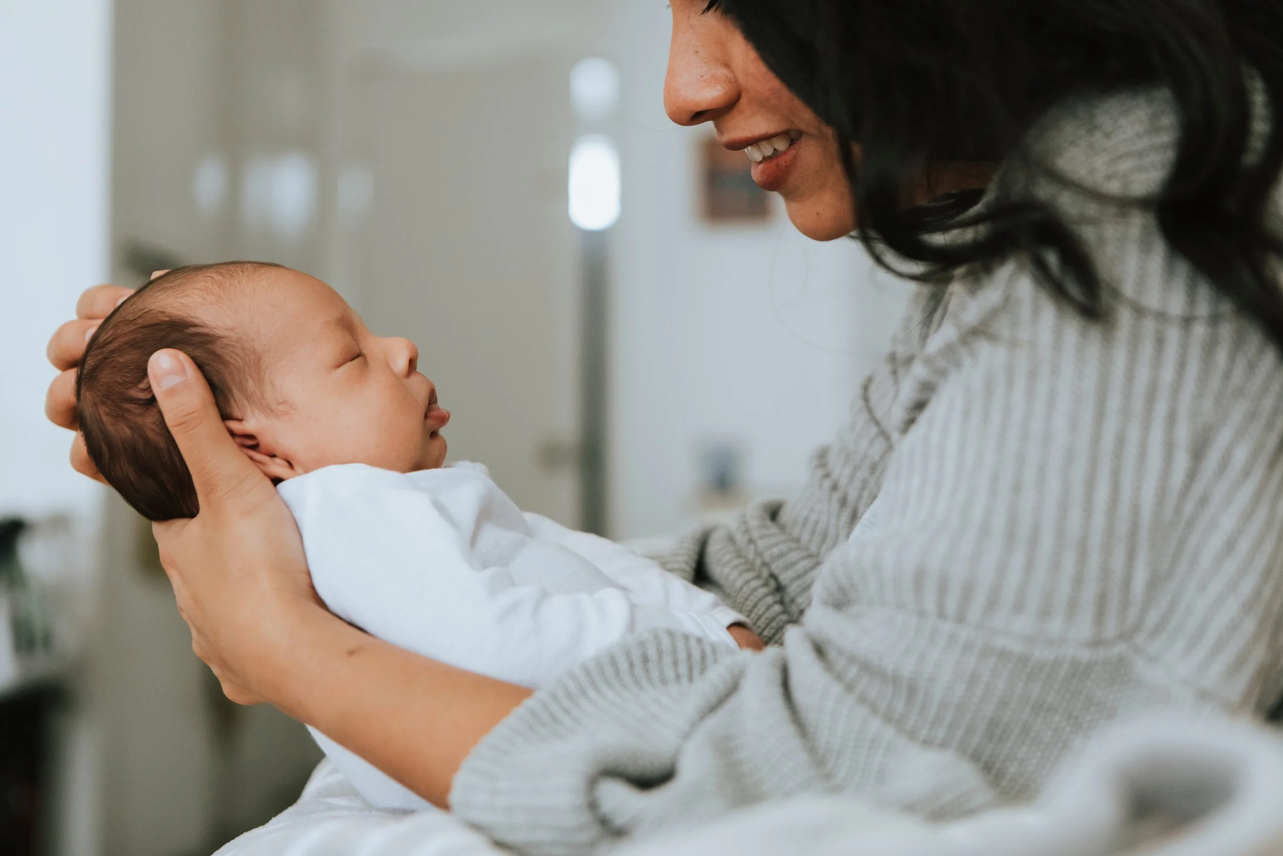A woman holding a sleeping baby close to her face, smiling softly. The baby is bundled in a white blanket or outfit and appears peaceful.