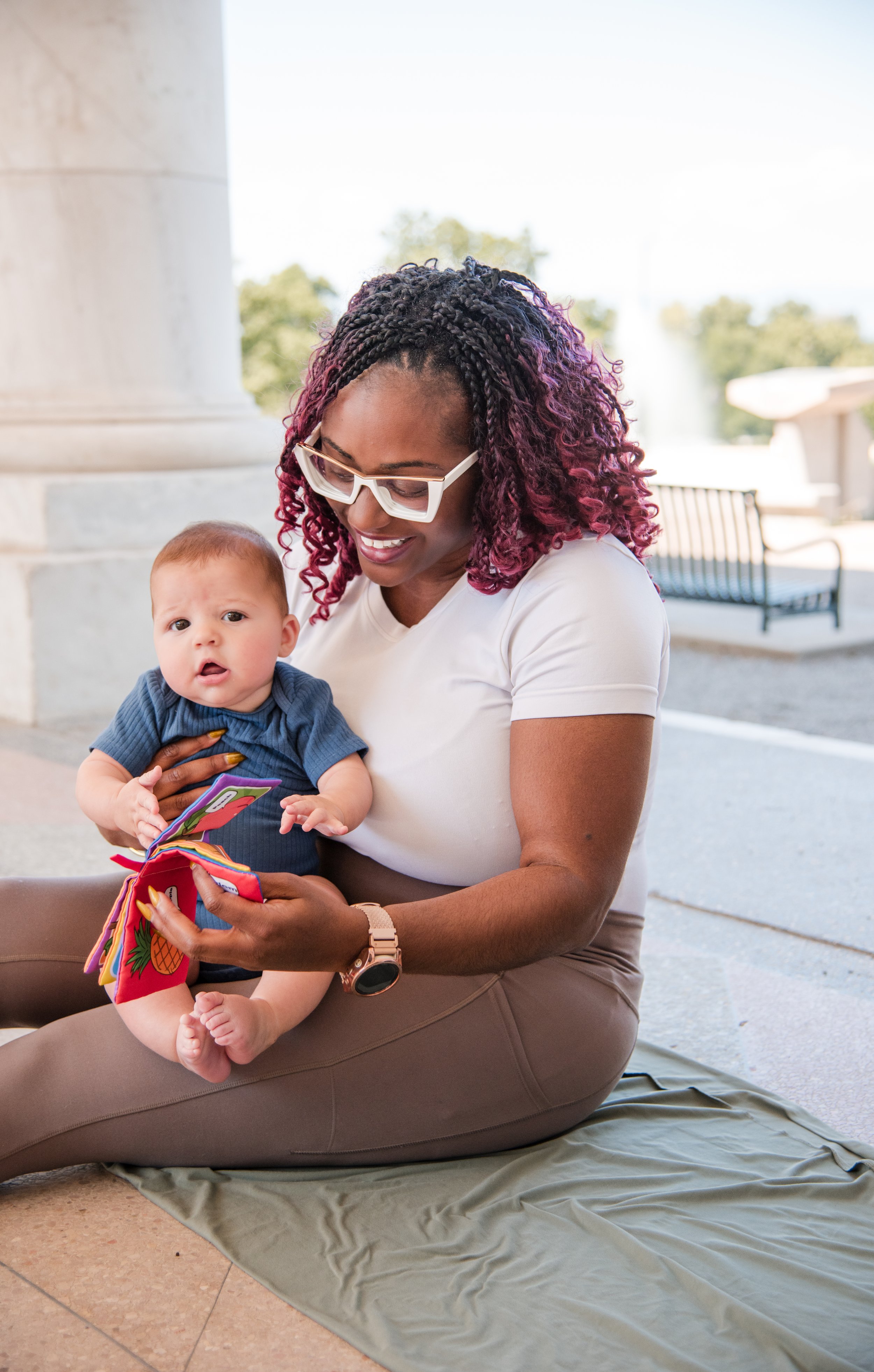 A woman sitting on a blanket outside, smiling at a baby she is holding, with a fountain and park benches in the background.