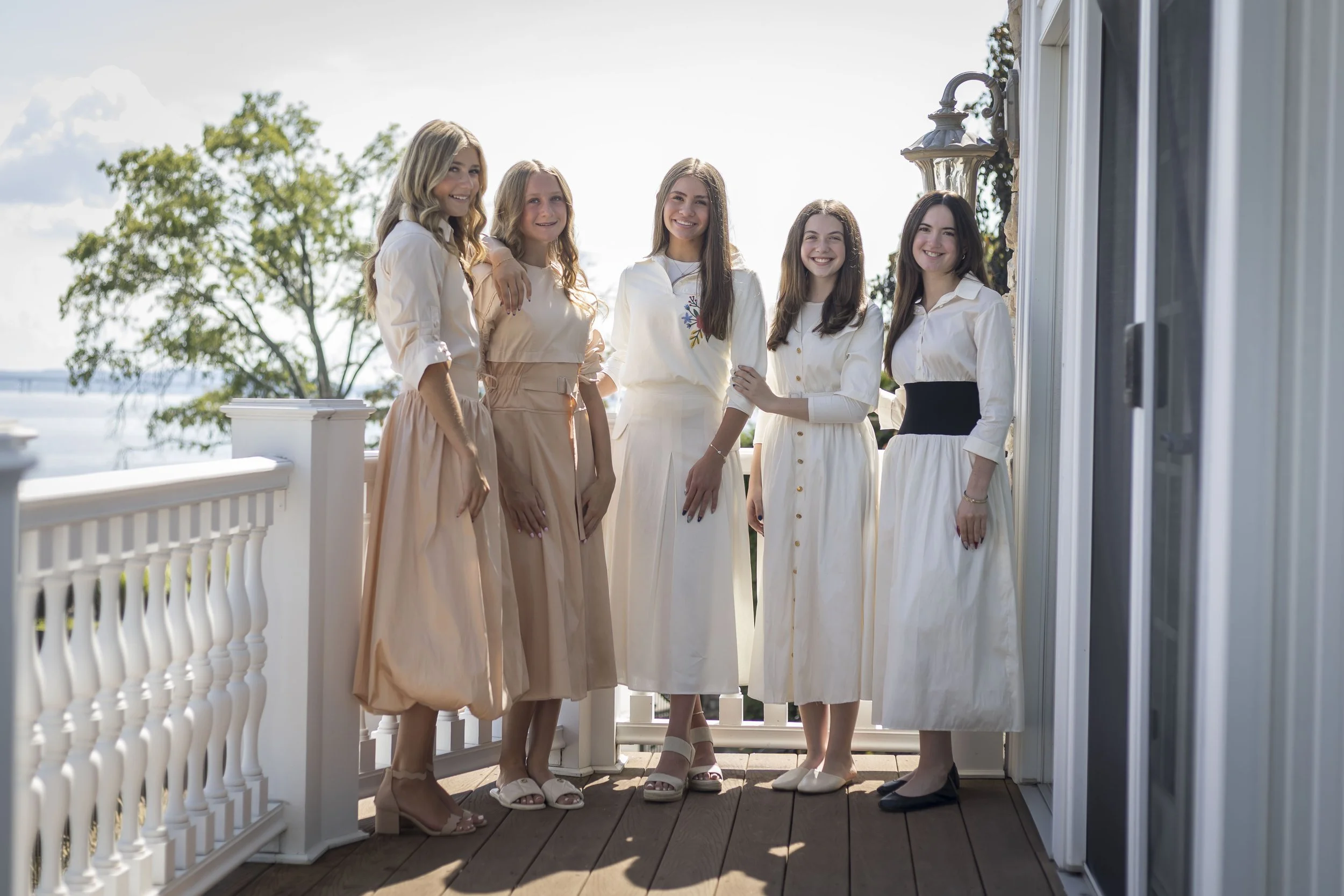 Five young women standing on a staircase dressed in white taffetta Shabbos dresses and black shoes, smiling for a group photo.