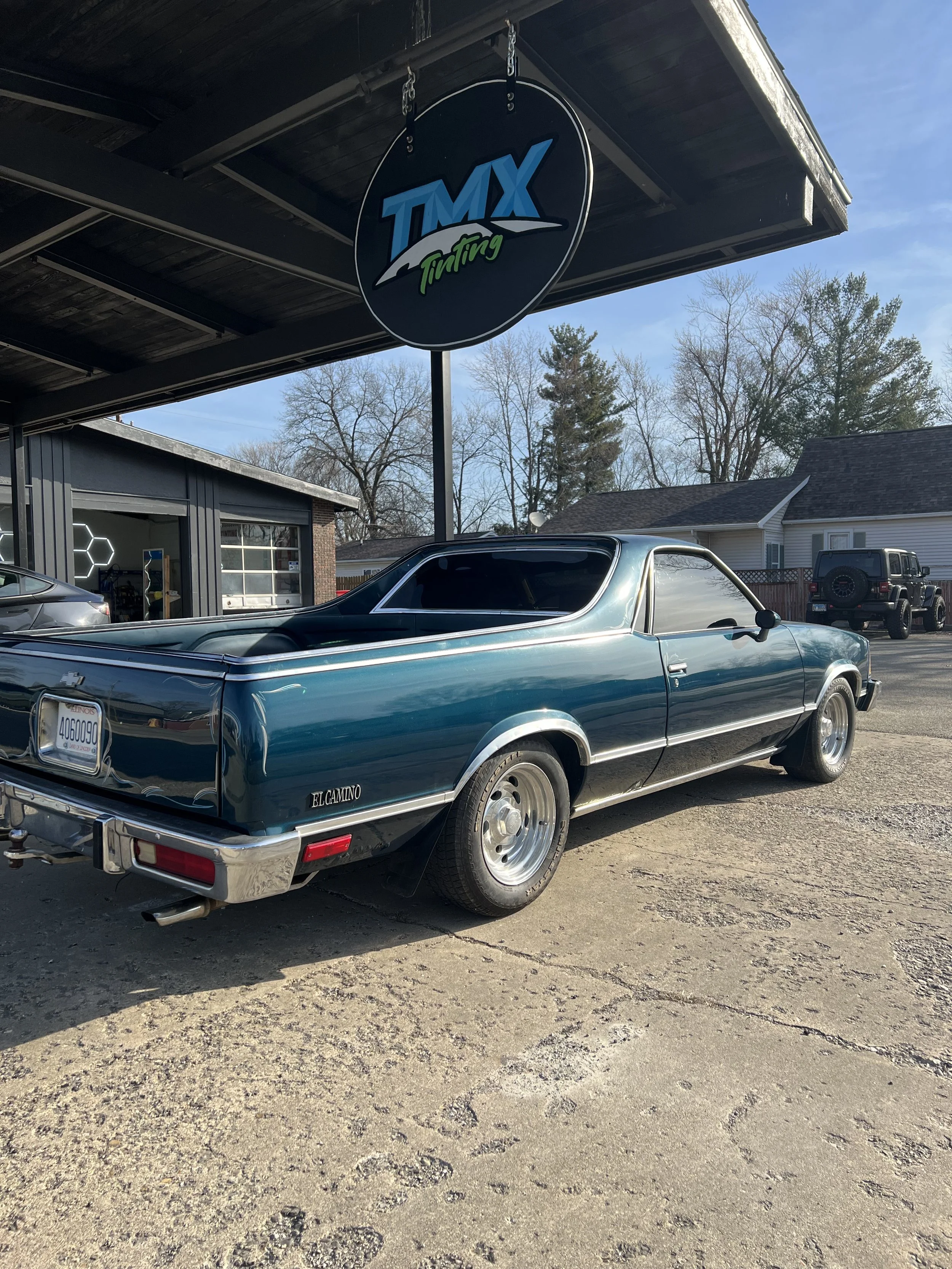 A vintage blue pickup truck parked in front of a tire shop with a sign that reads 'TMX Tuning' on a sunny day.