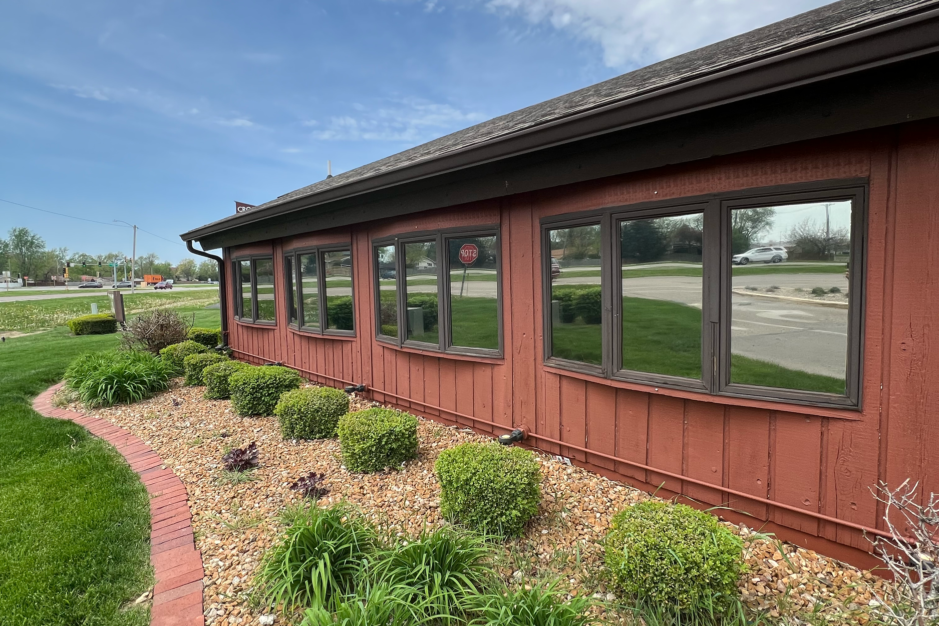 Exterior of a red wooden building with multiple windows, landscaped with green bushes and small plants, on a sidewalk with a street and moving vehicles in the background.