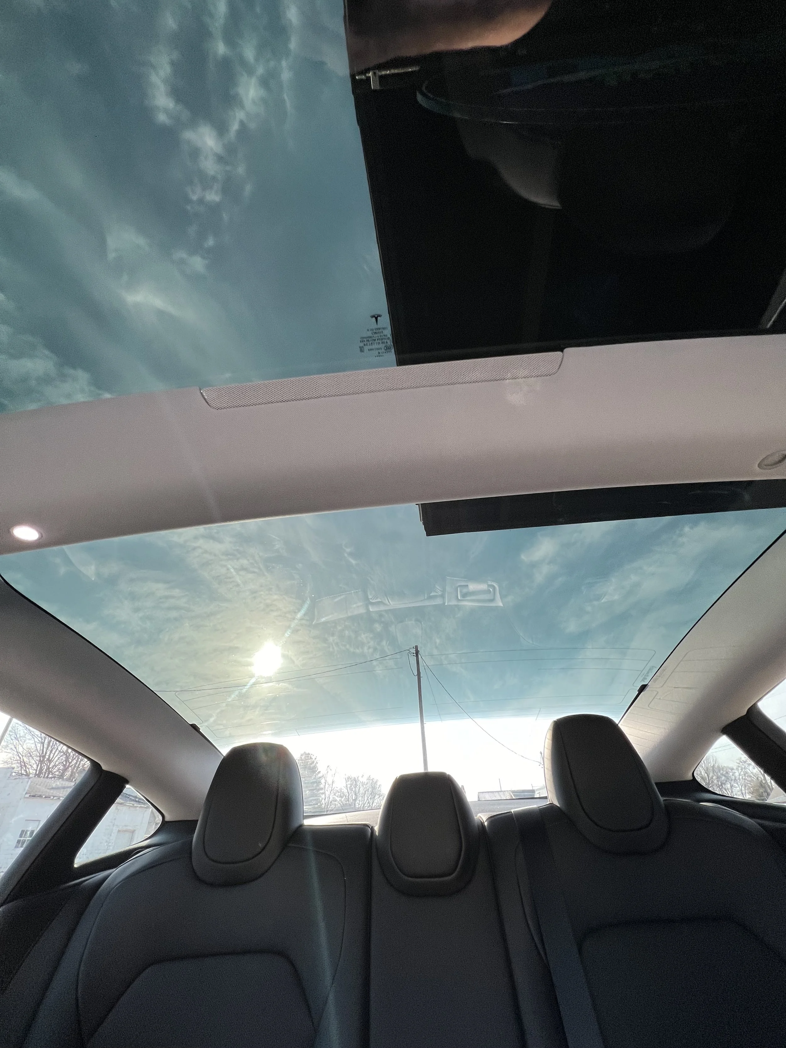 View of the interior of a Tesla electric vehicle showing the large glass roof, the back seats, and the sky outside with clouds and the sun.