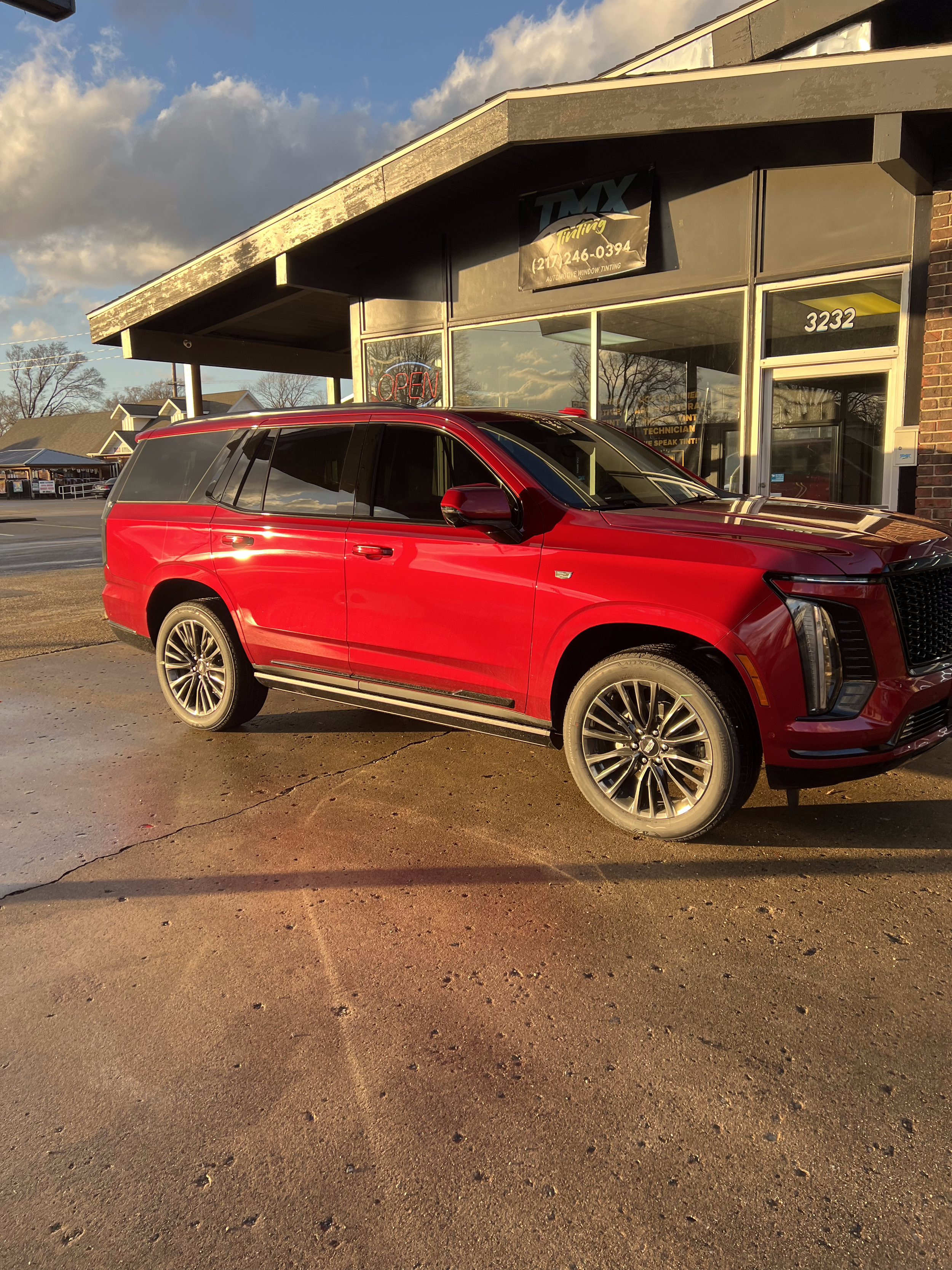 Red SUV parked outside car repair shop with large windows, sign reading 'OPEN', and building number 3232, in late afternoon sunlight.