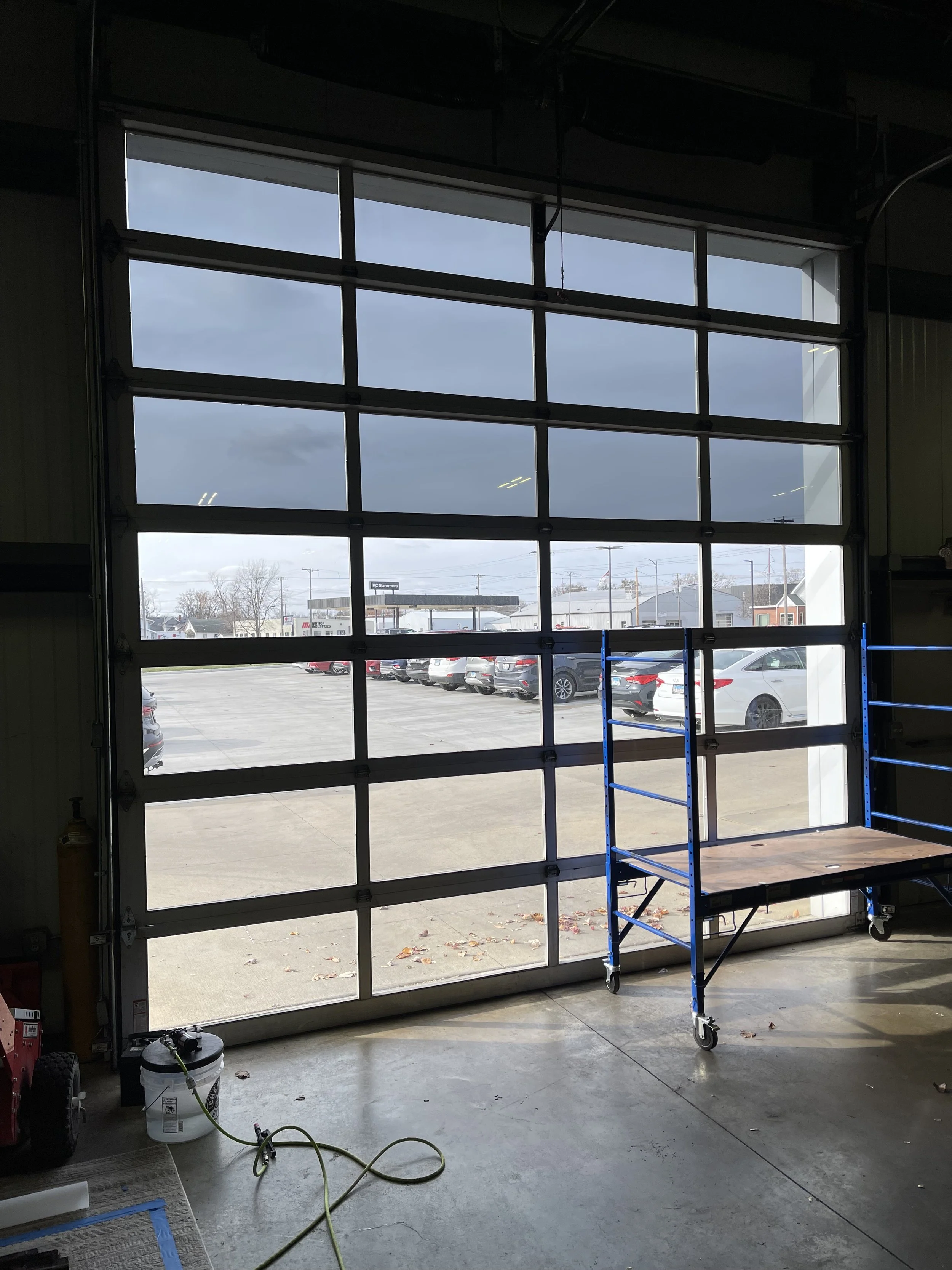 View through an open garage door showing a parking lot with several cars, a gas station, and buildings in the distance. Inside the garage, there is a blue tool cart and some equipment.