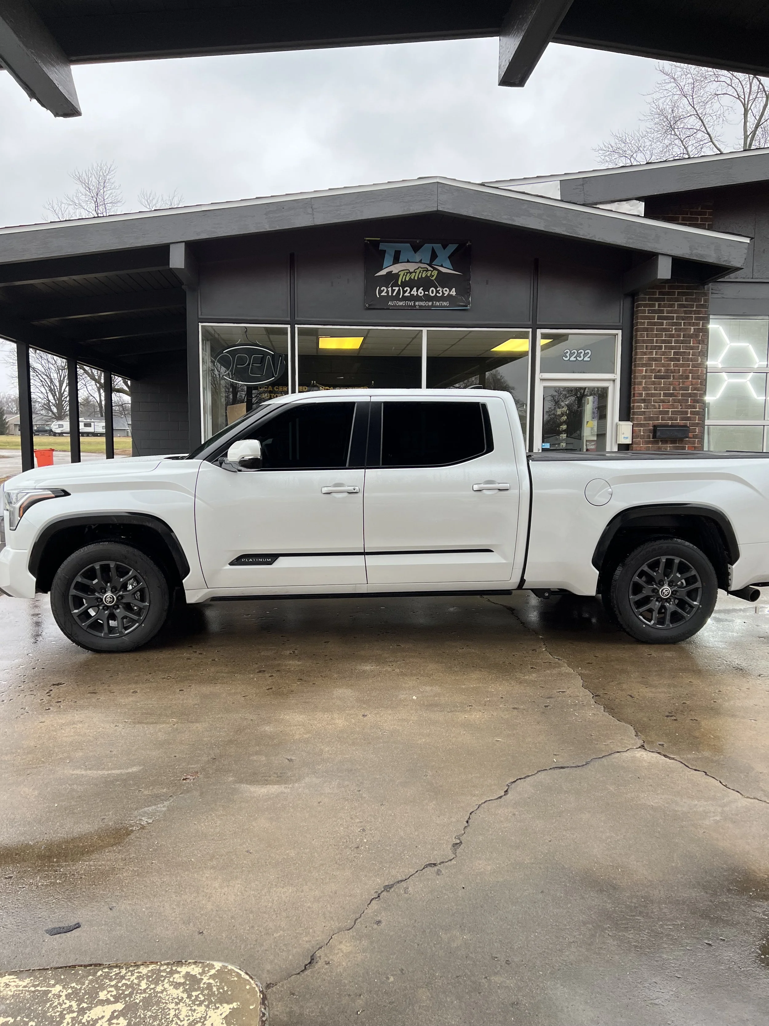 White pickup truck parked in front of an automotive tinting shop with a black sign and large glass windows.