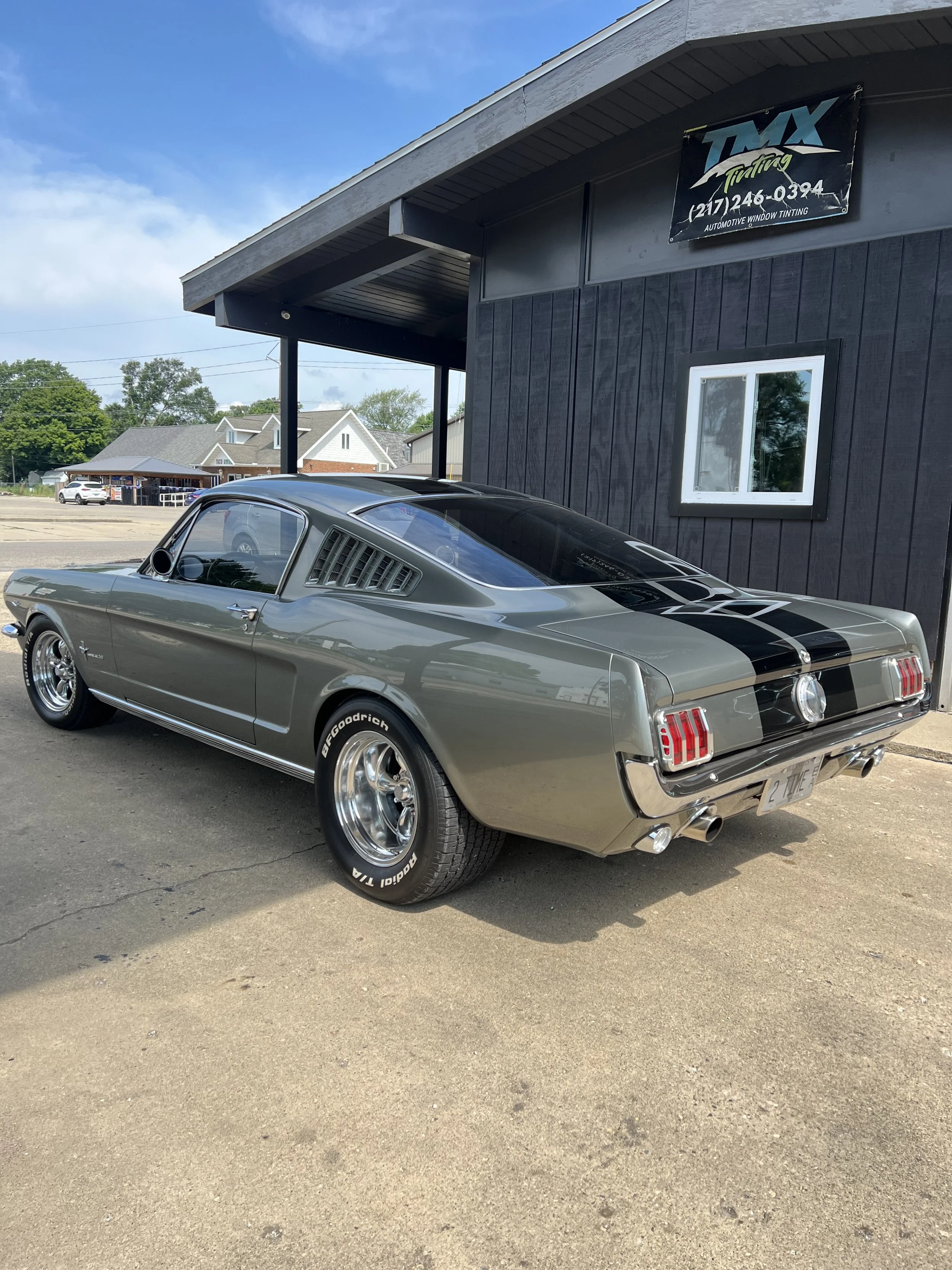 A classic silver Ford Mustang with black racing stripes parked outside a black building with a sign that reads 'TMX Tinting'.