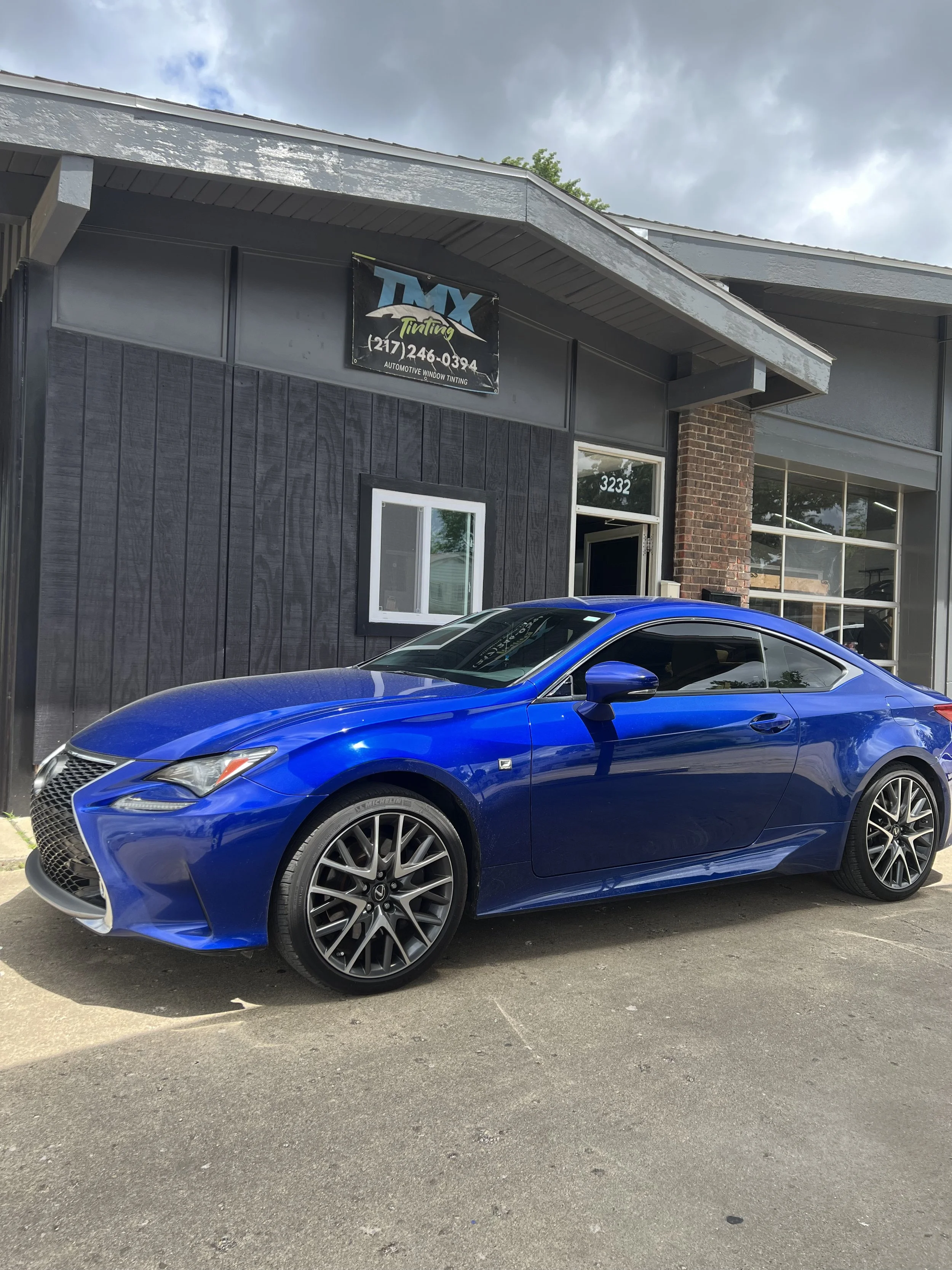 Blue Lexus RC coupe parked outside a black and gray automotive window tinting shop.