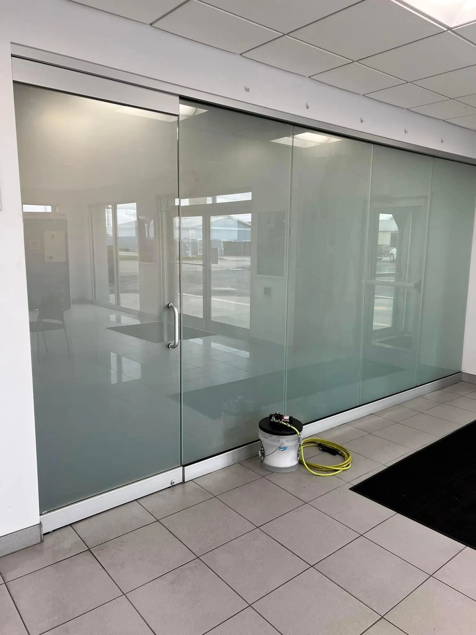 Empty office or commercial space with glass wall and door, tiled floor, and a bucket connected to cleaning equipment in the corner.