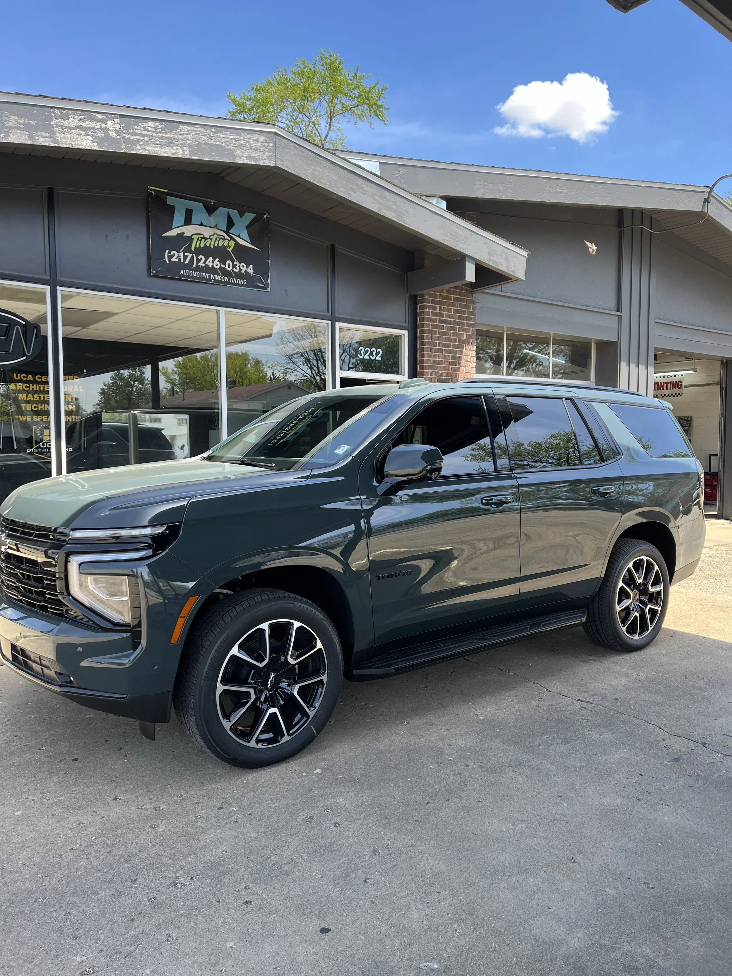 A black SUV parked outside an automotive window tinting shop with a sign that reads 'TMX Tinting' and a phone number. The shop is in a brick and gray building with large glass windows, and there are green trees and a blue sky with clouds visible in t