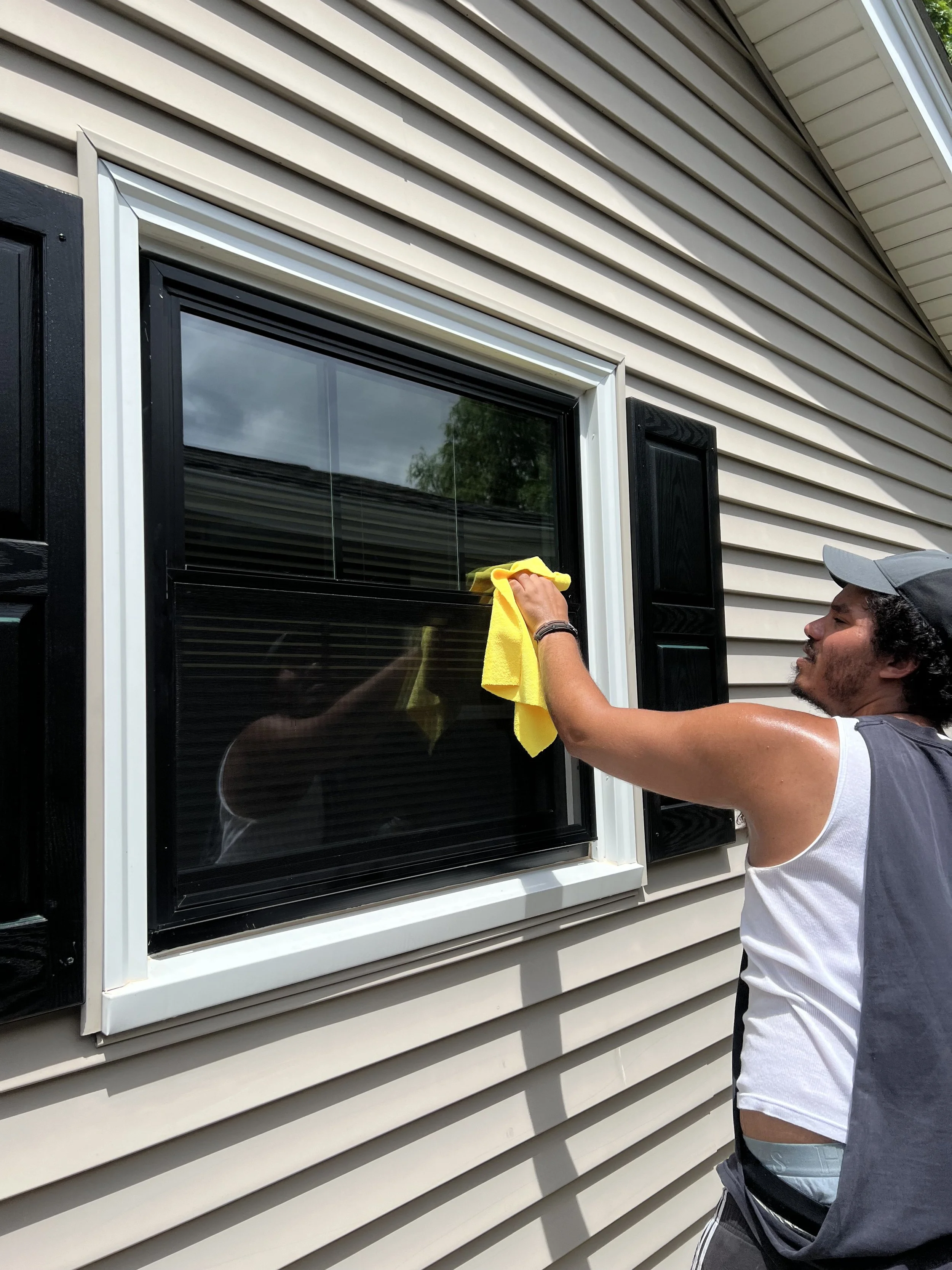 A man cleaning a window with a yellow cloth outside a house with beige vinyl siding and black window shutters.