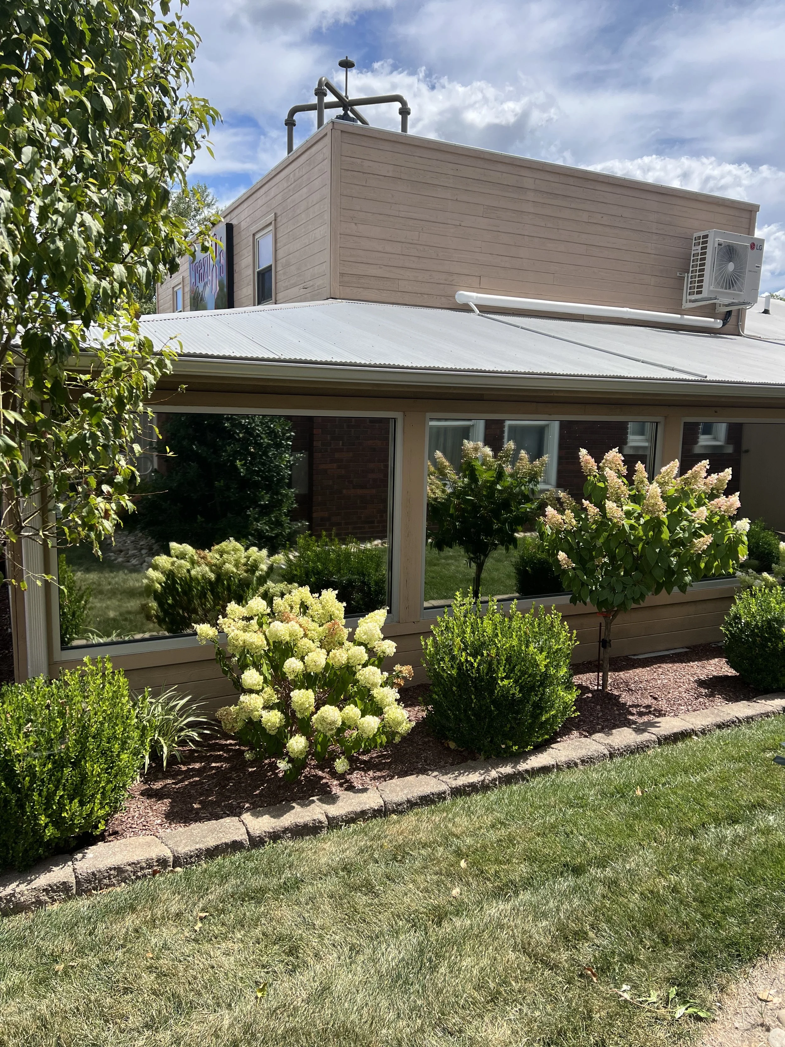 View of a building with a garden in front, featuring blooming bushes, green shrubs, and grass, under a partly cloudy sky.