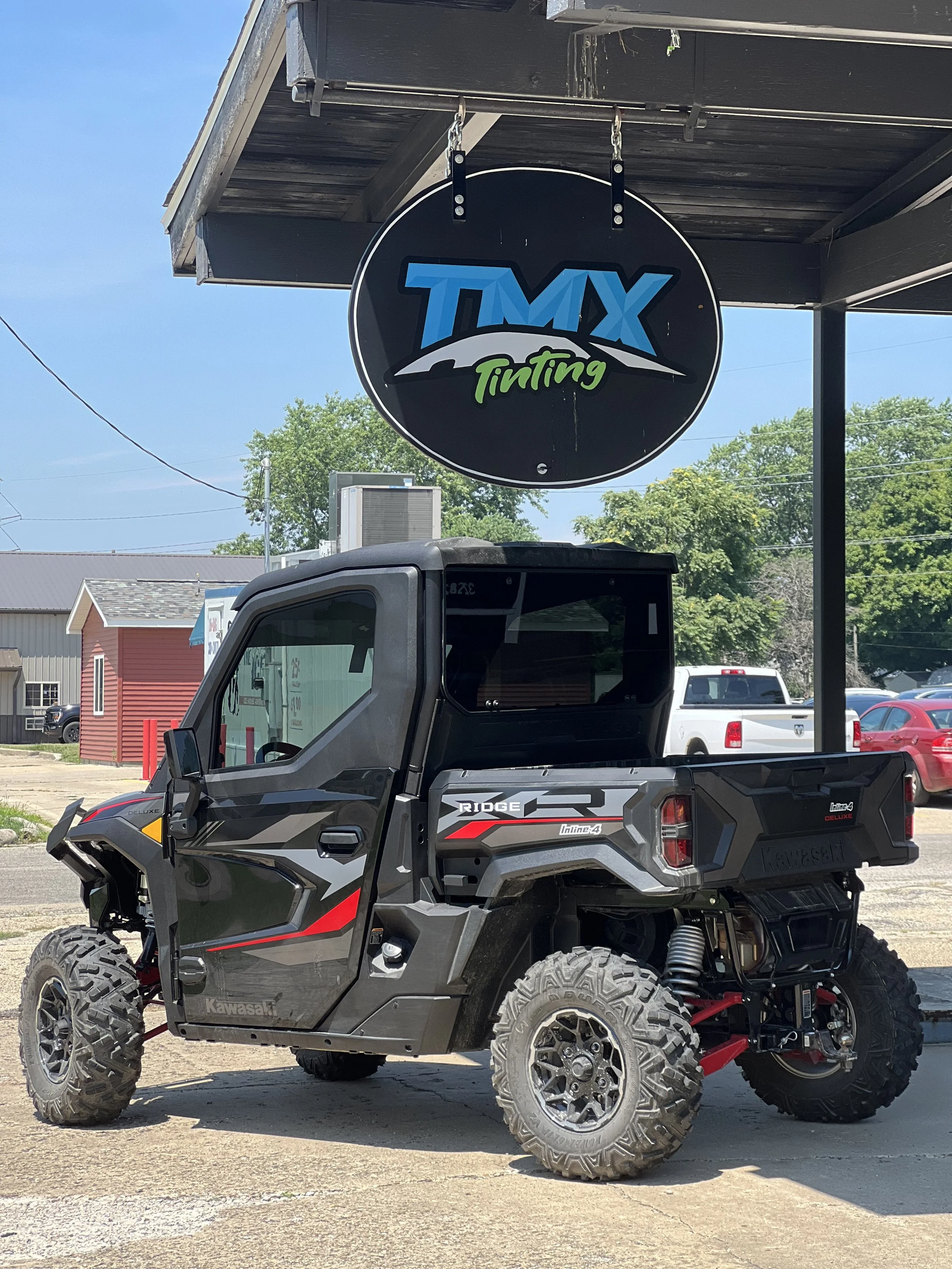 A Kawasaki all-terrain vehicle parked outside a tinting shop called TMX Tinting, with a sign hanging above. The vehicle has large off-road tires and a matte black and gray body with red accents.