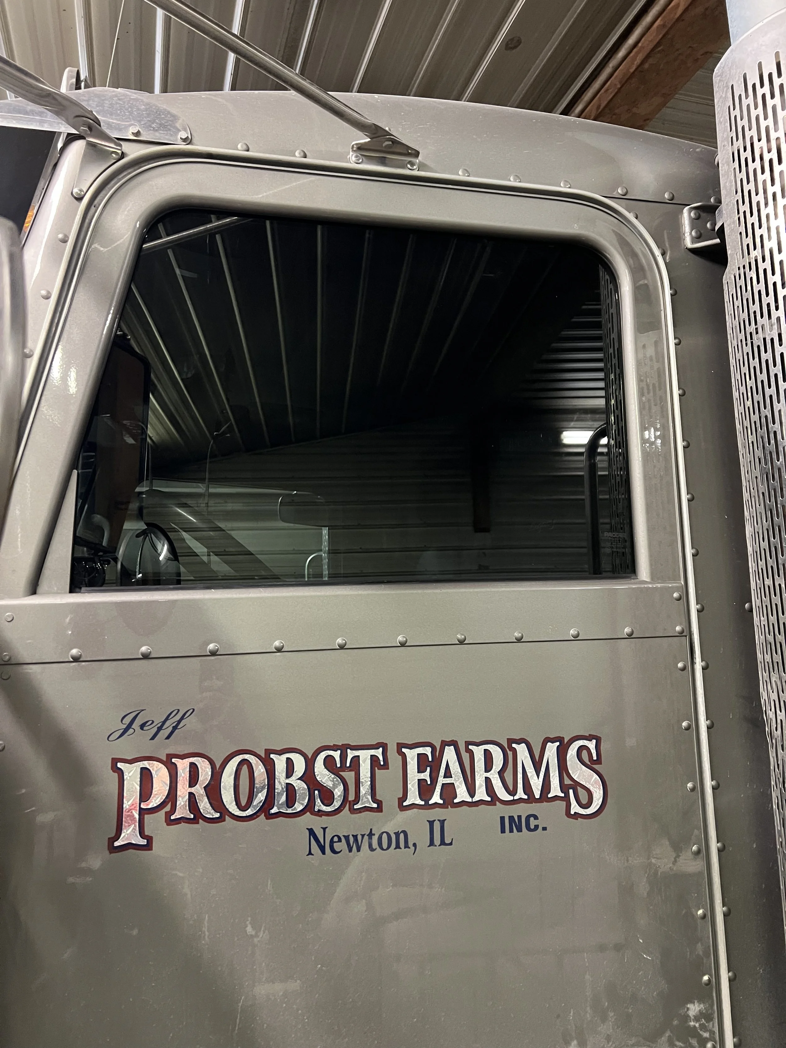 Close-up of a truck cab with a silver exterior, showing the window and part of the door. The door has a logo that reads "Jeff PROBST FARMS INC. Newton, IL" in stylized font.