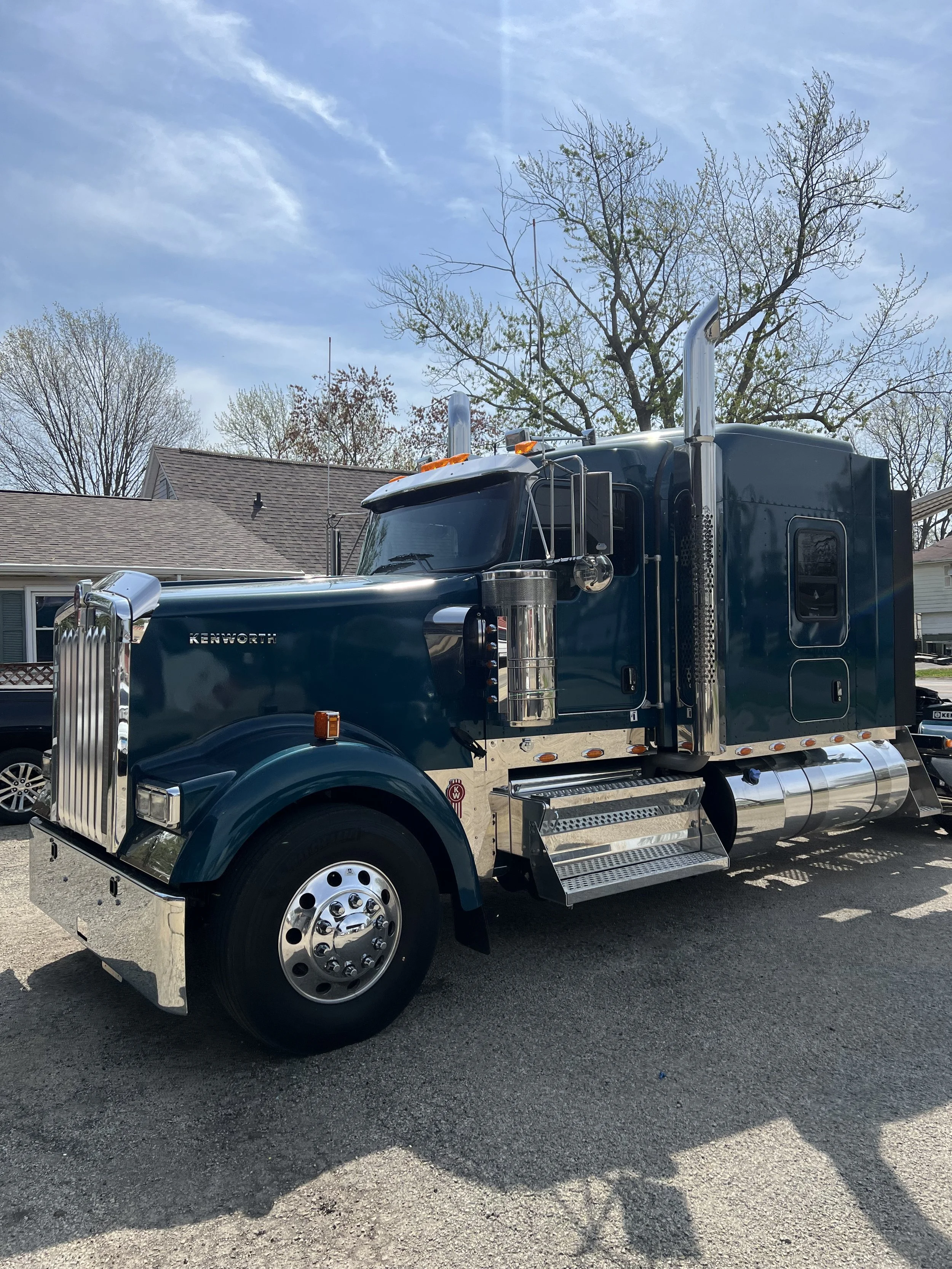 Blue semi-truck with chrome details parked on a street with houses and leafless trees under a partly cloudy sky.