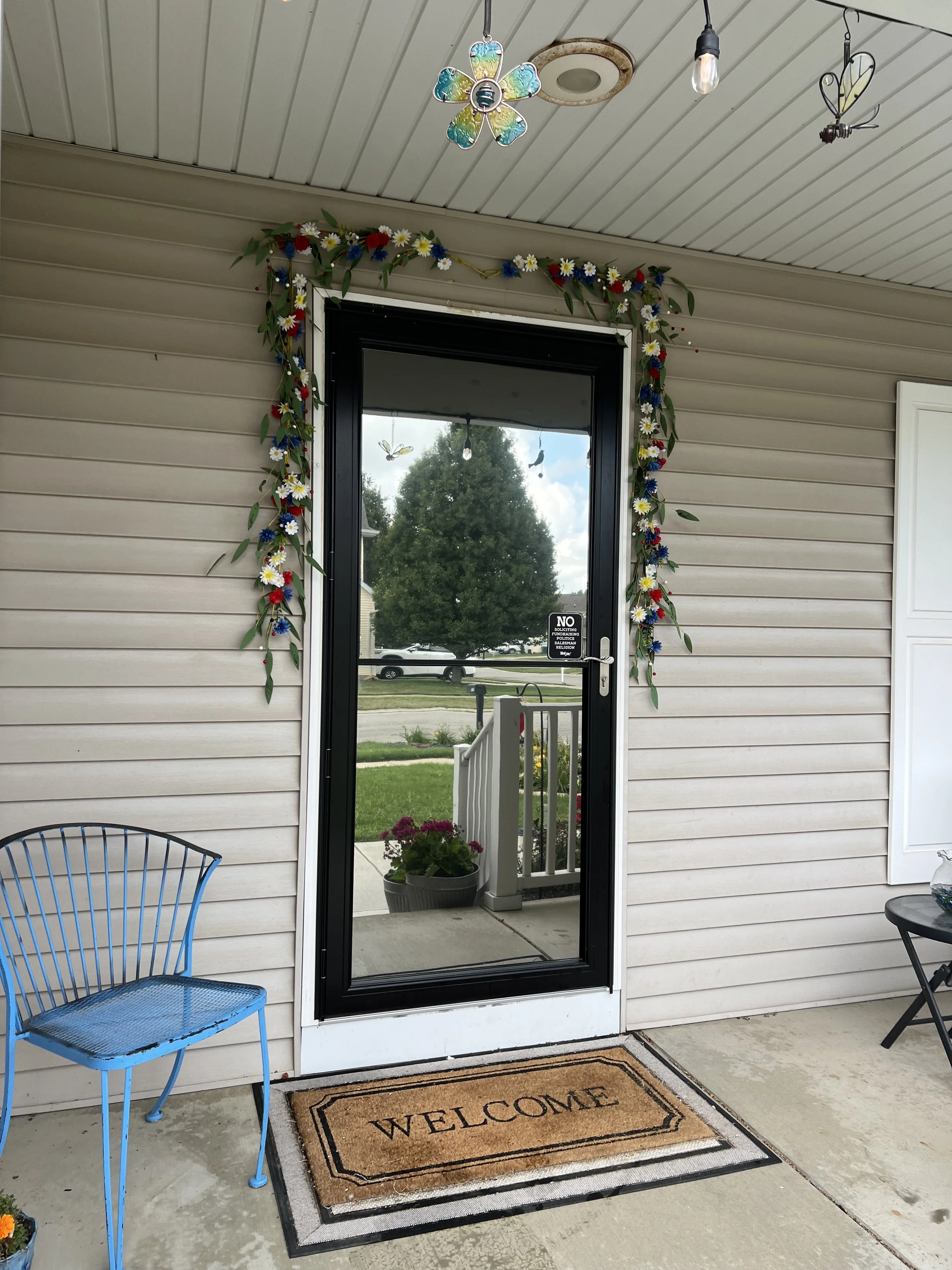 A porch with a glass door surrounded by a floral wreath, a welcome mat, and outdoor furniture, including a blue metal chair and a small black table with a glass pitcher, under a ceiling with hanging stained glass flower and butterfly decorations.