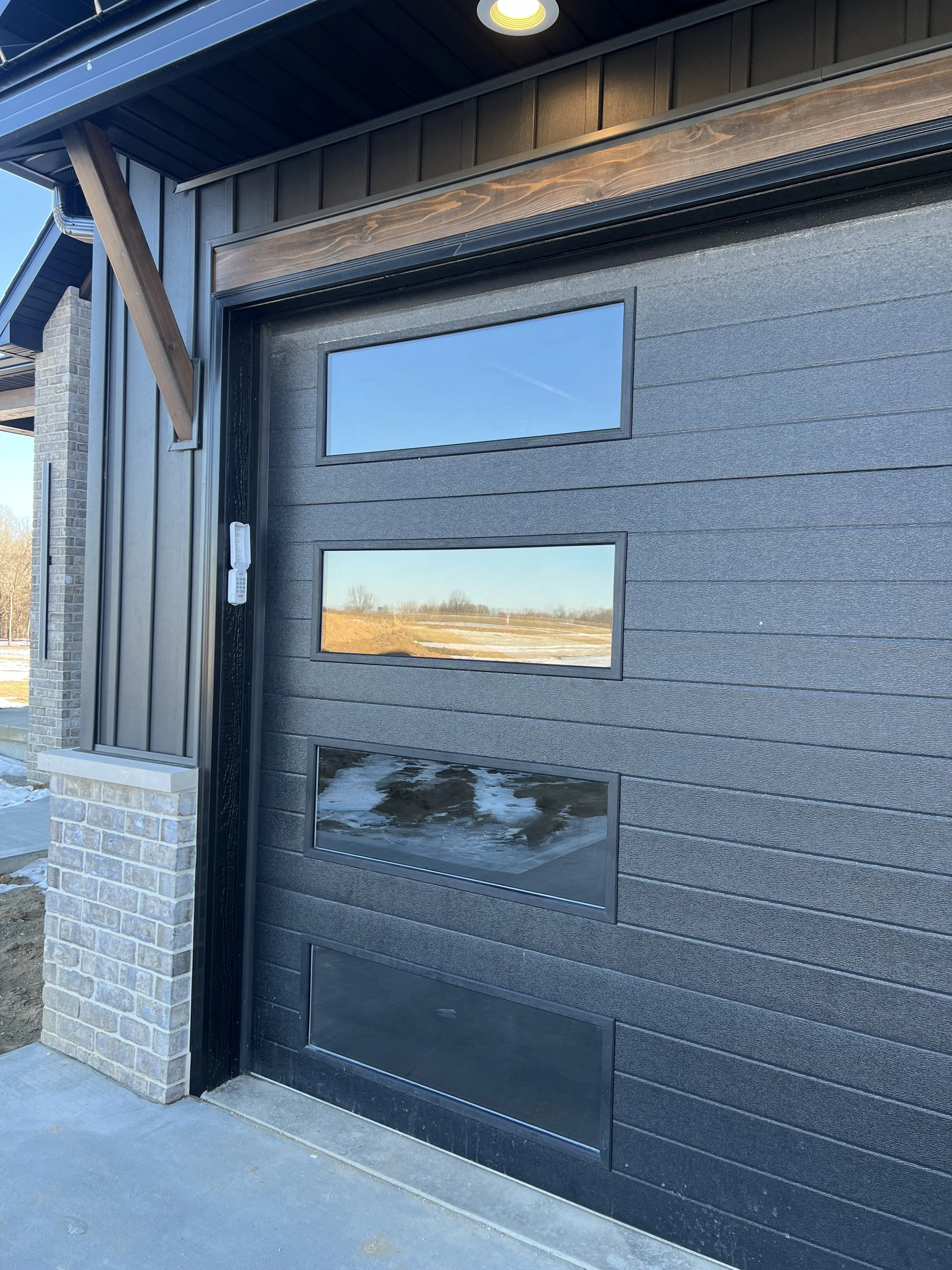 Close-up of a modern black garage door with four rectangular windows, reflecting the blue sky and outdoor landscape, with brick and wood siding on the building.