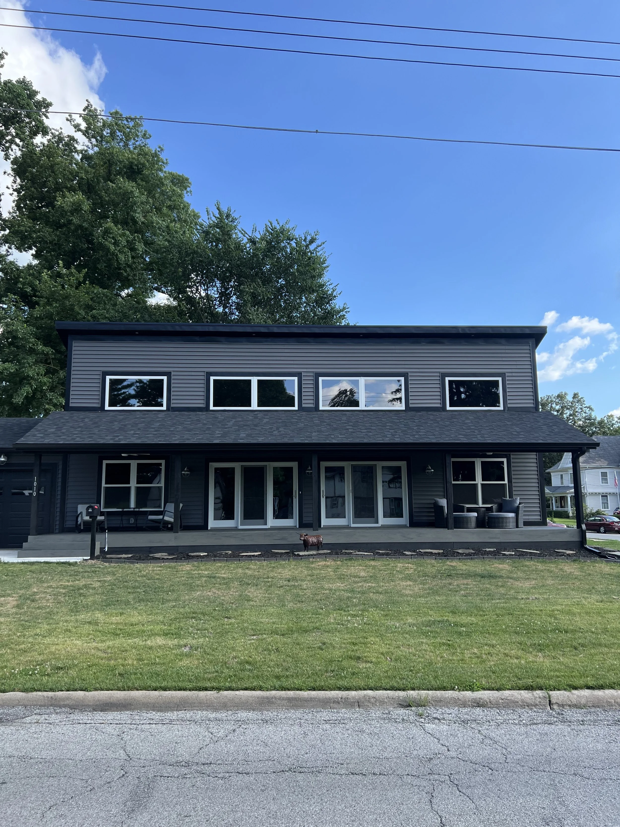 A modern two-story house with a dark exterior, large windows, a covered front porch, and outdoor seating, situated on a grassy lawn with a paved street in front. Trees are visible behind the house and the sky is partly cloudy.
