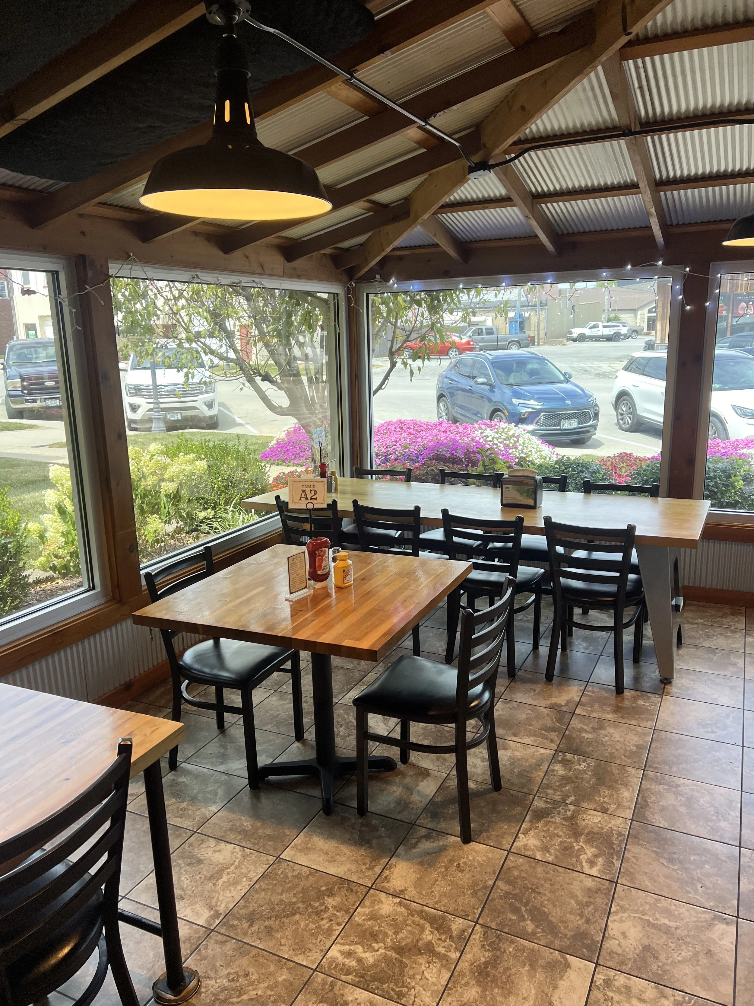 Inside a cozy restaurant with large windows overlooking a parking lot and colorful flowers, featuring wooden tables, black chairs, and warm lighting.
