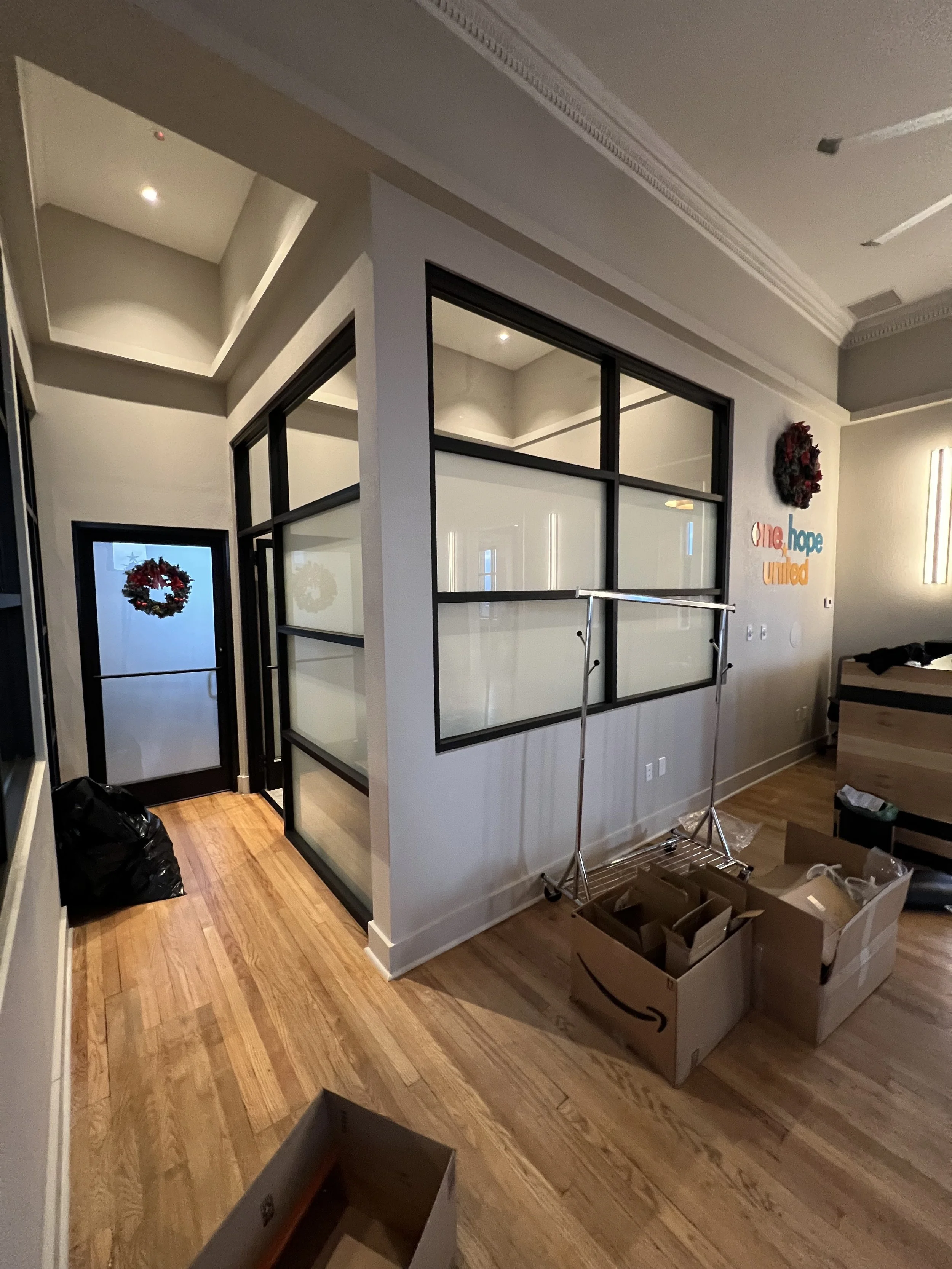 Interior of a house with wooden floors, decorated for Christmas with wreaths and a sign 'one hope united'. There are moving boxes and a clothing rack in the room, with a black trash bag in the corner and a glass partition wall.