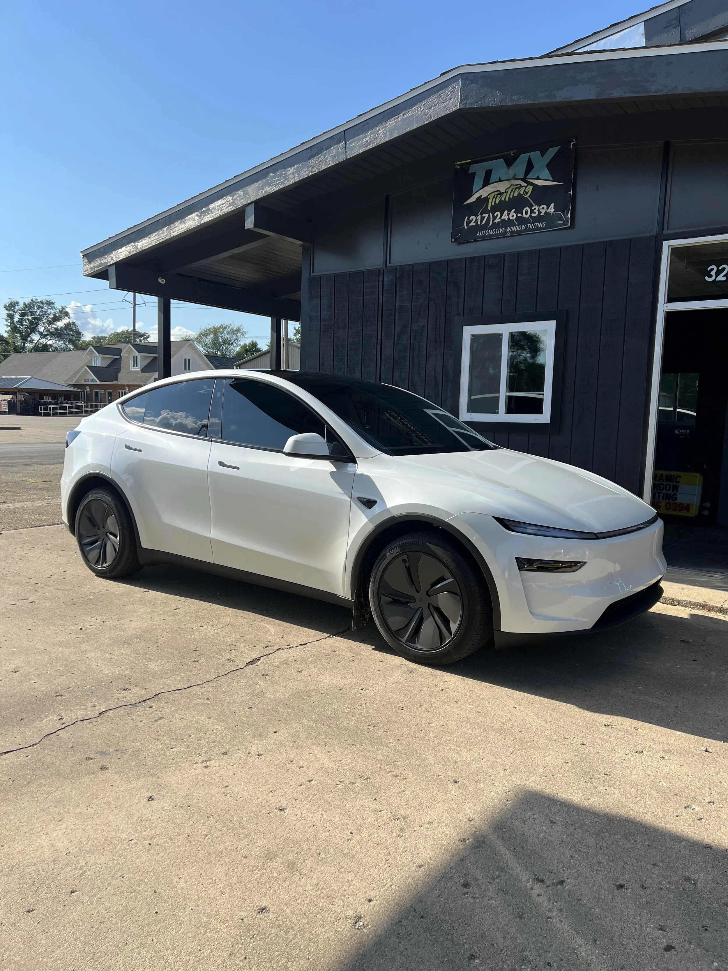White Tesla Model Y parked outside a dark-colored automotive window tinting shop, with a sign above that reads 'TMX Tinting' and a phone number.