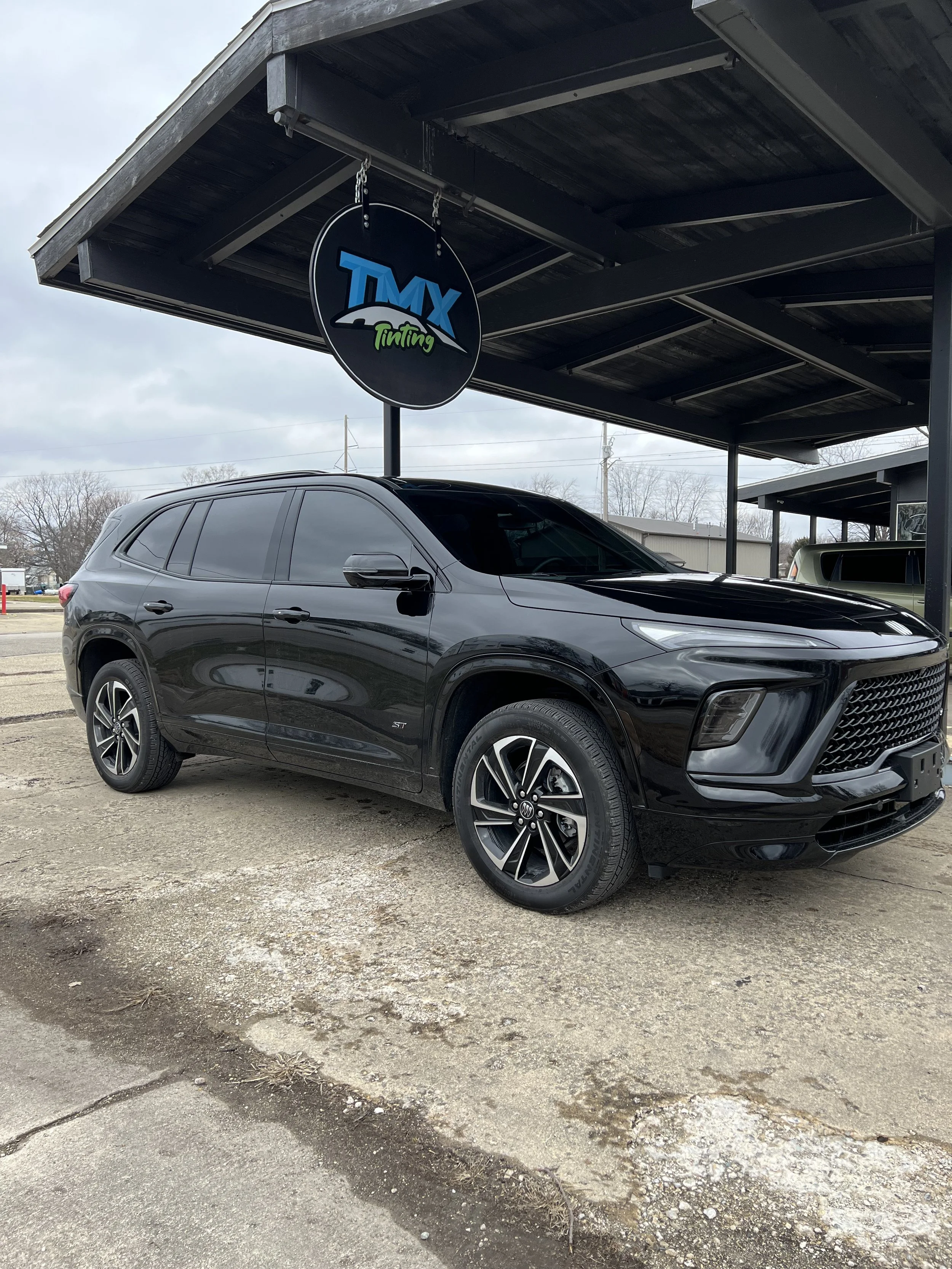 Black SUV parked under a carport with a sign that reads "TMX Tinting" hanging from the roof.