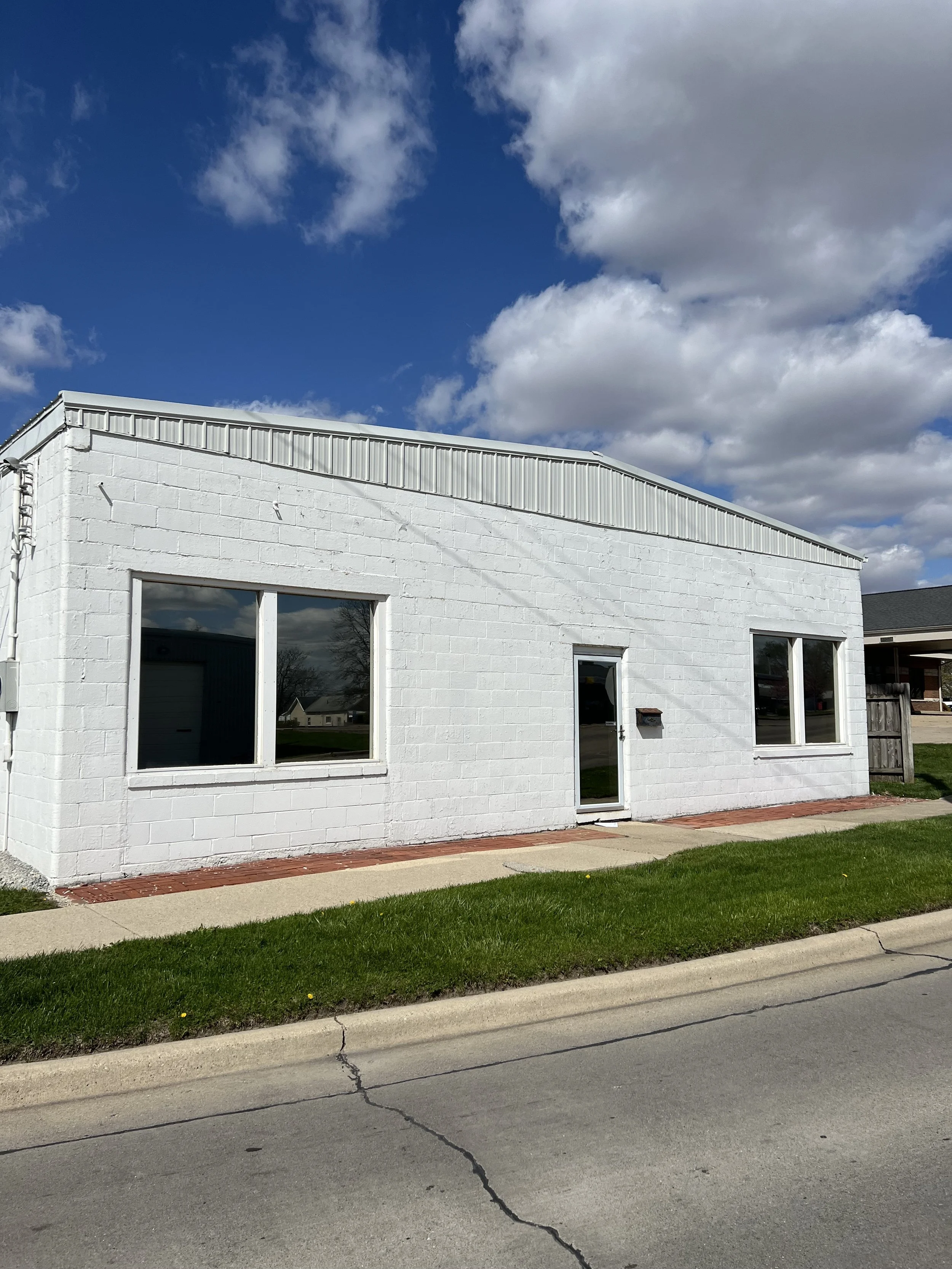 A white brick building with two large windows and a door, under a partly cloudy sky with green grass and sidewalk in front.