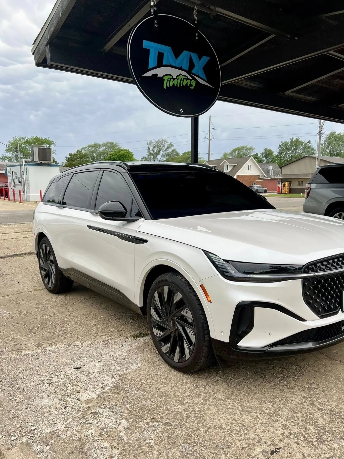 A white SUV with tinted windows parked outside a tinting shop under a black awning with the shop's circular sign hanging above. The sign reads 'TMX Tinting' with a stylized mountain and sky background. In the background, there are houses, trees, and other vehicles.