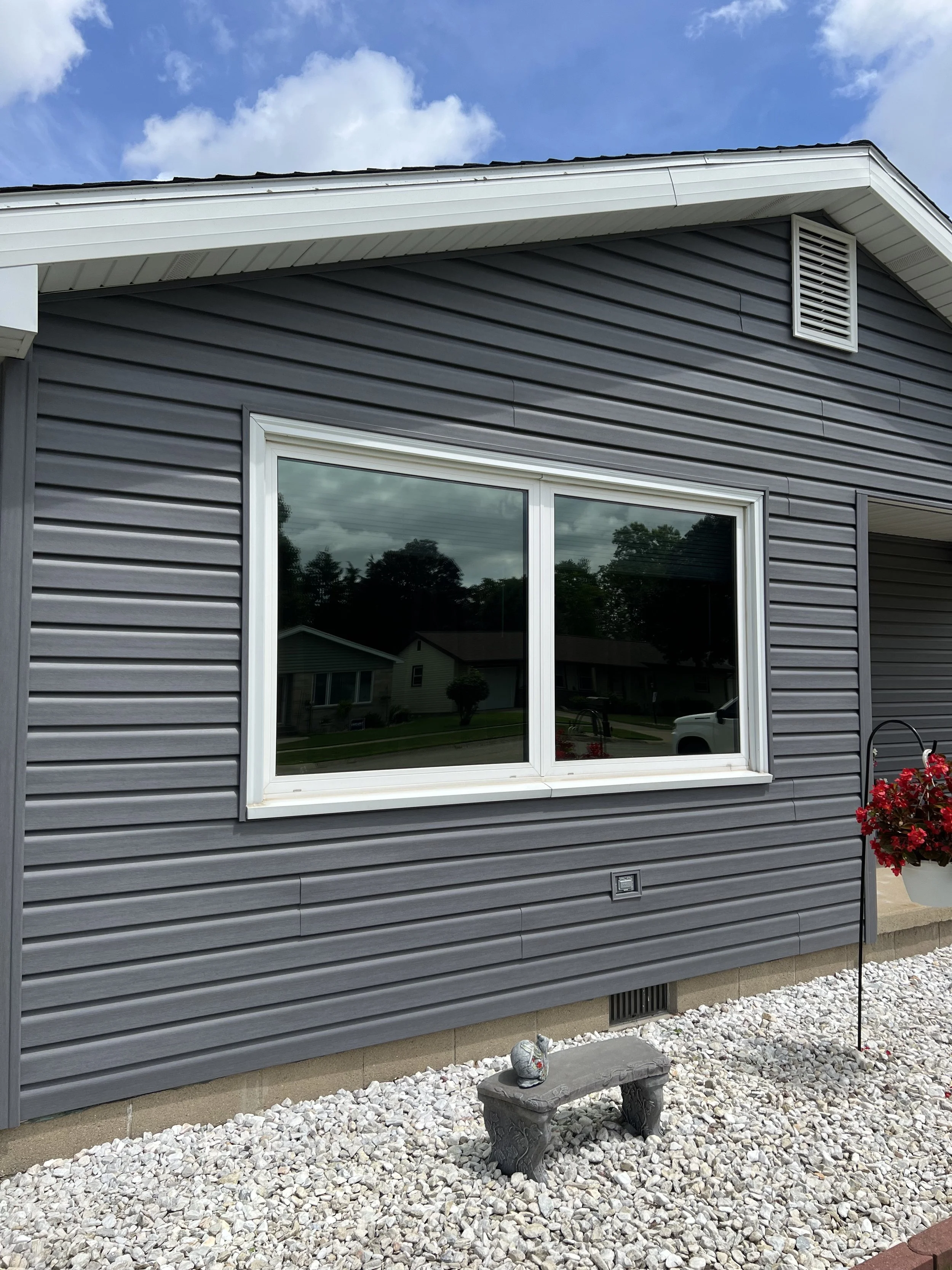 The exterior of a house with gray siding, a white-framed window, a vent near the roof, and a hanging flower basket; a small stone bench with a decorative egg on top, gravel ground, and some plants and houses reflected in the window.