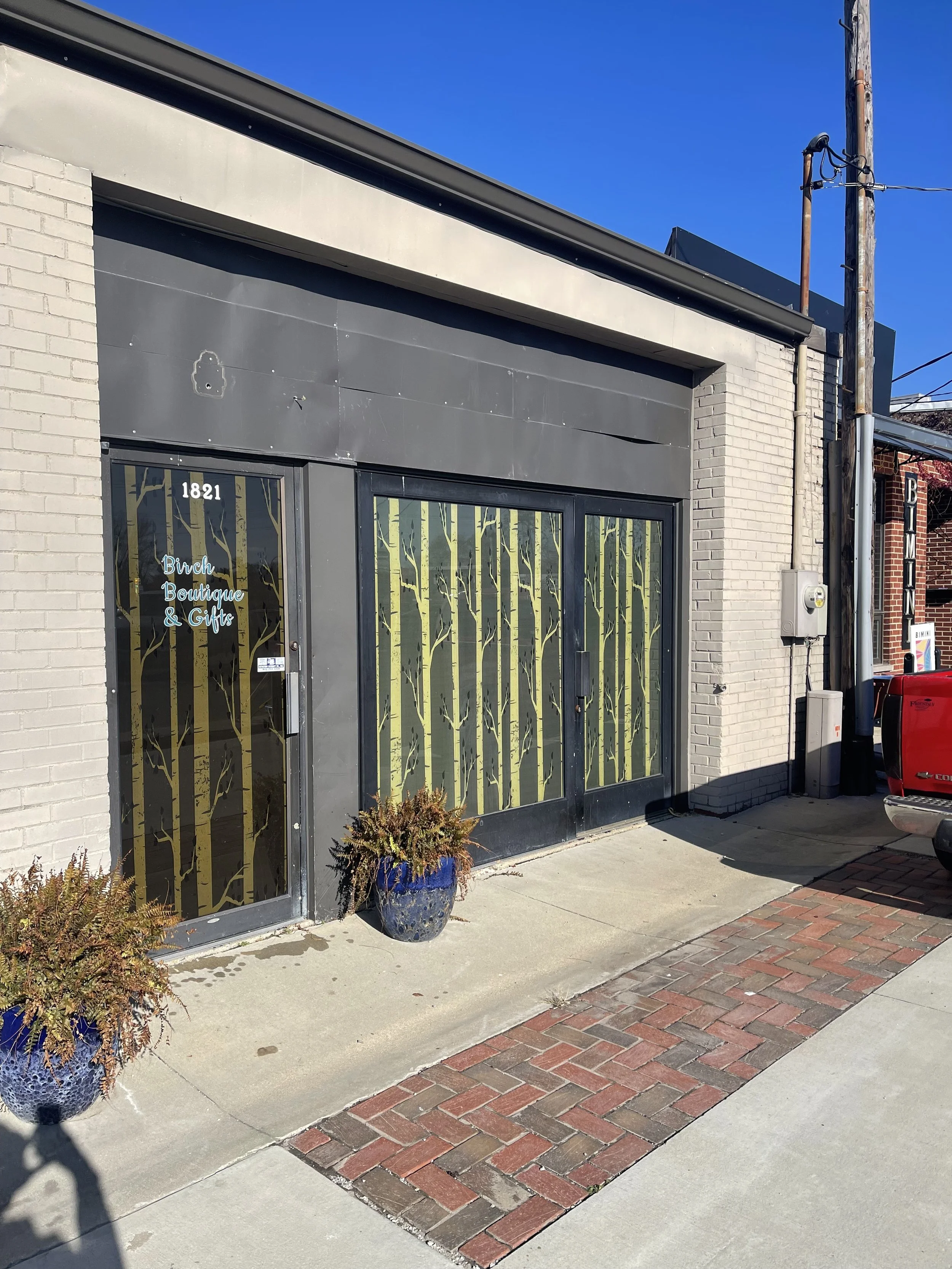 The storefront of Birch Boutique & Gifts, located at 1821, with large windows decorated with a birch tree design, two potted plants outside, and a sidewalk with brick and concrete paving.