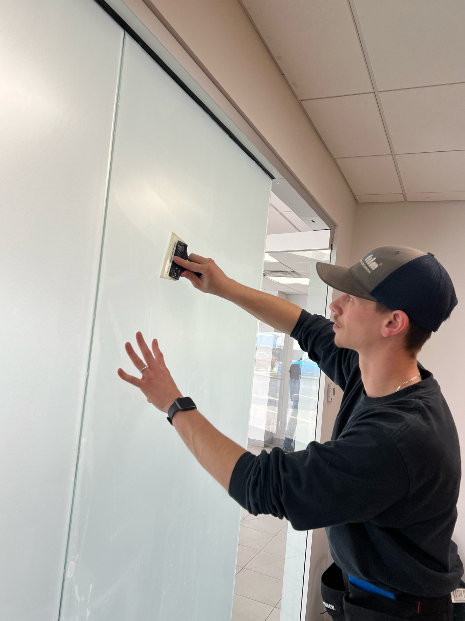 A man cleaning a glass wall with a squeegee, wearing a black long-sleeve shirt, a baseball cap, and an Apple Watch.