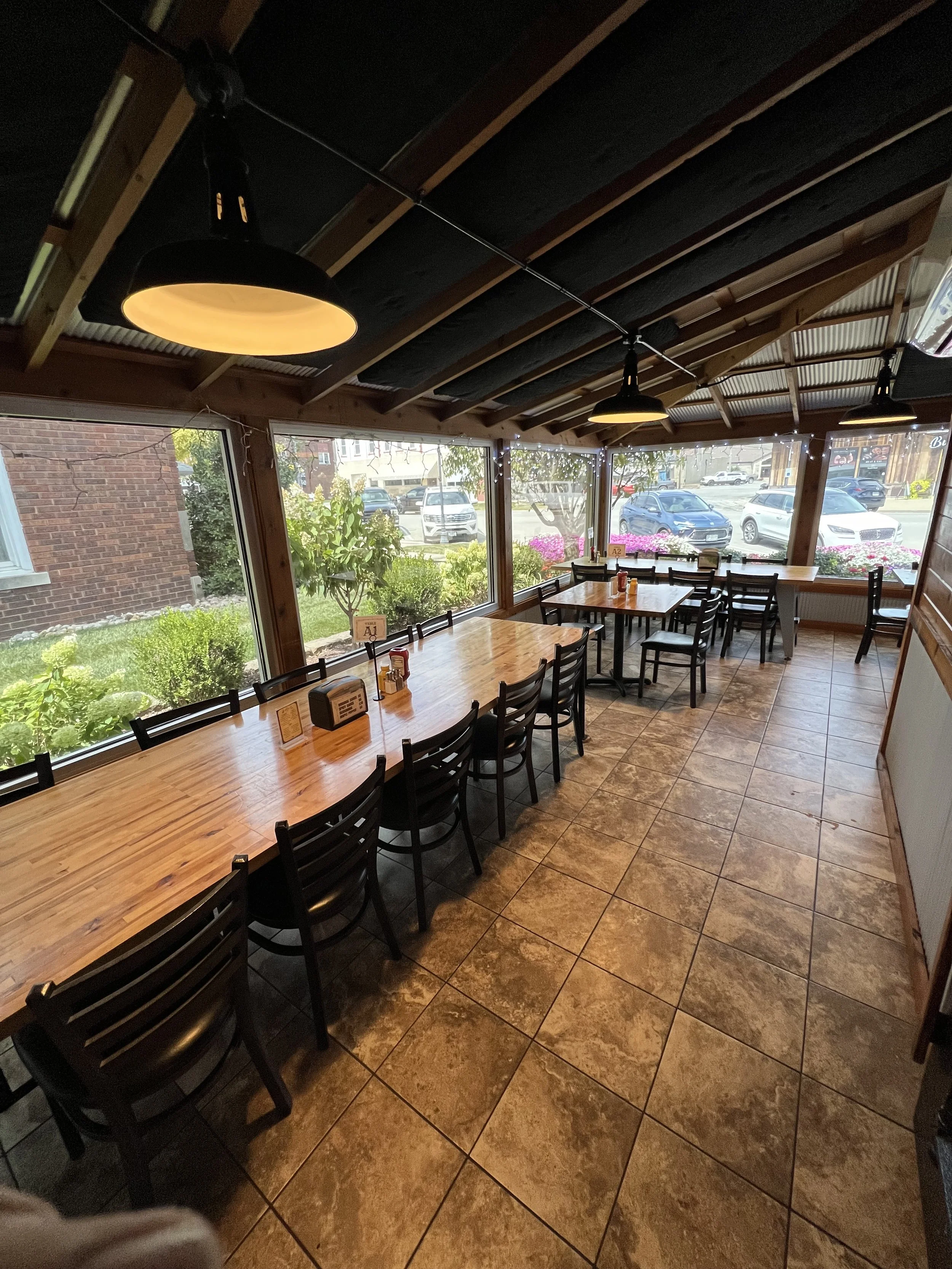 Interior of a restaurant with a long wooden table, black chairs, large windows, and ceiling lights.