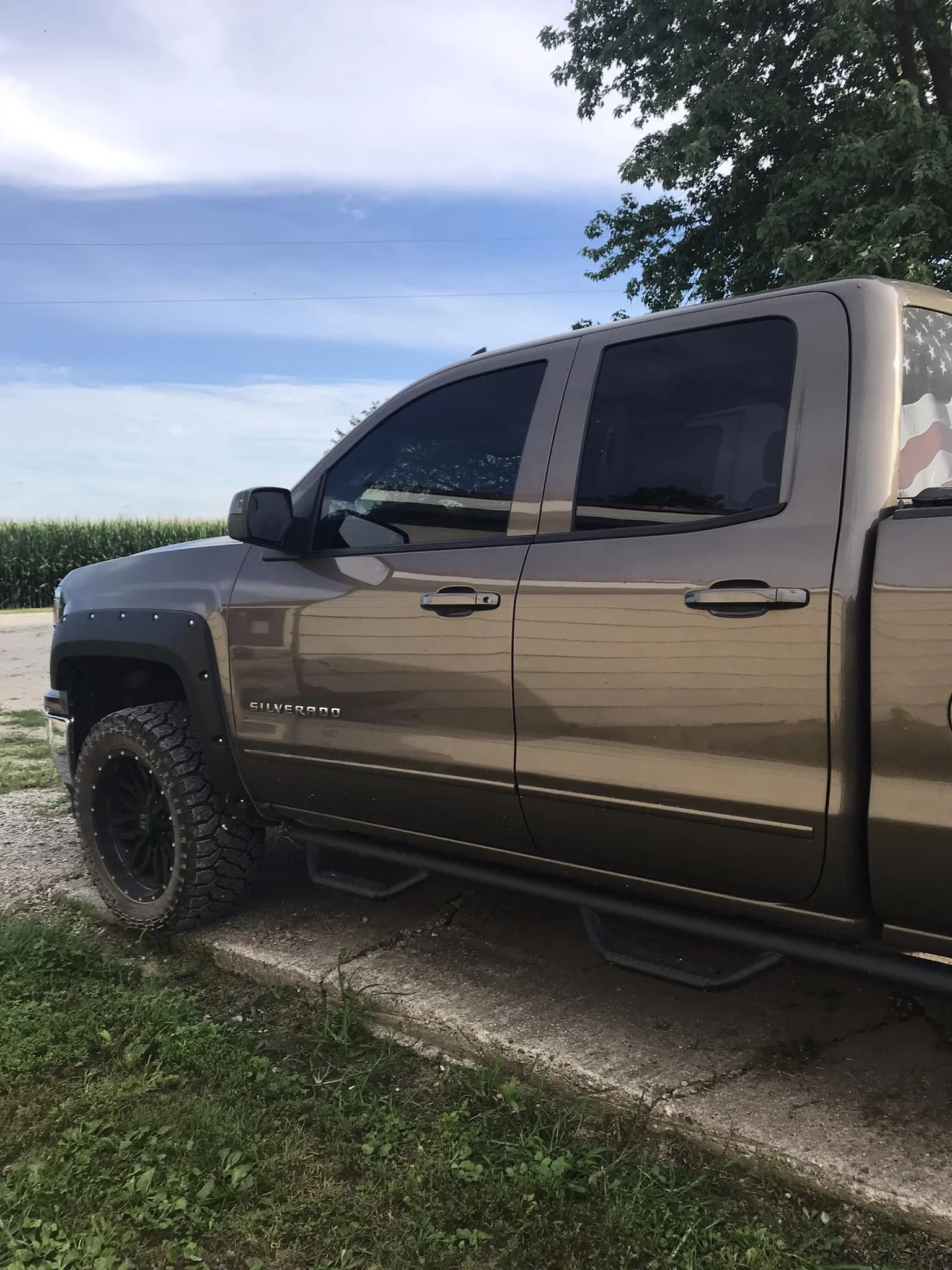 A metallic brown Chevrolet Silverado pickup truck parked on a concrete slab beside a grassy area with a field of crops and trees in the background, under a partly cloudy sky.