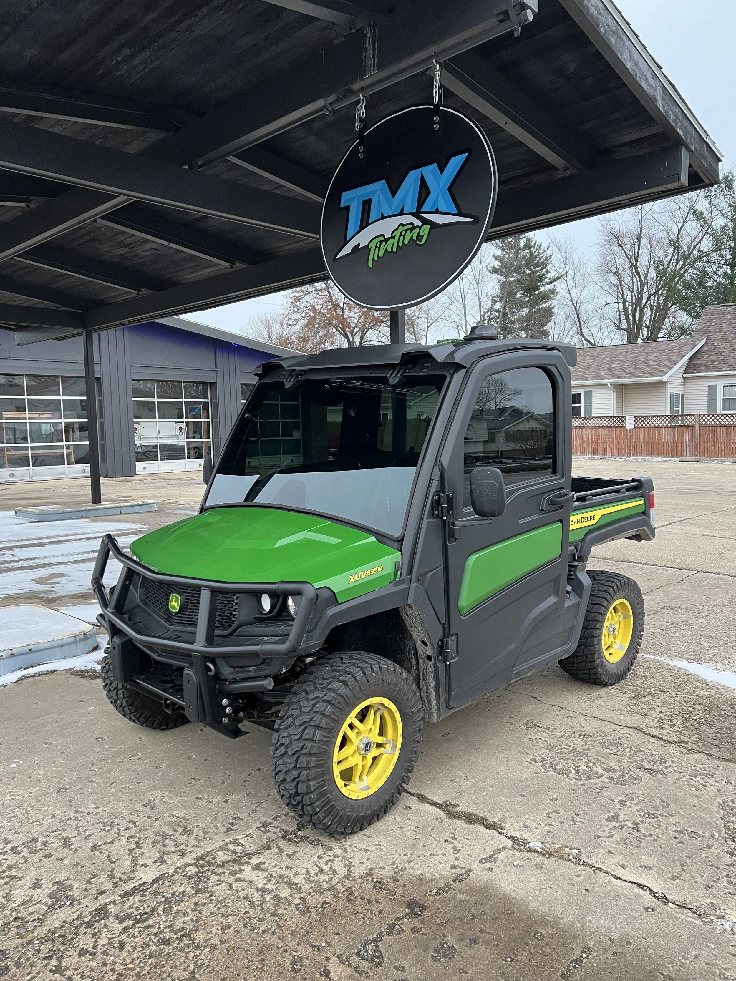 Green and black John Deere utility vehicle parked under a covered area with a sign for TMX Tunting overhead.