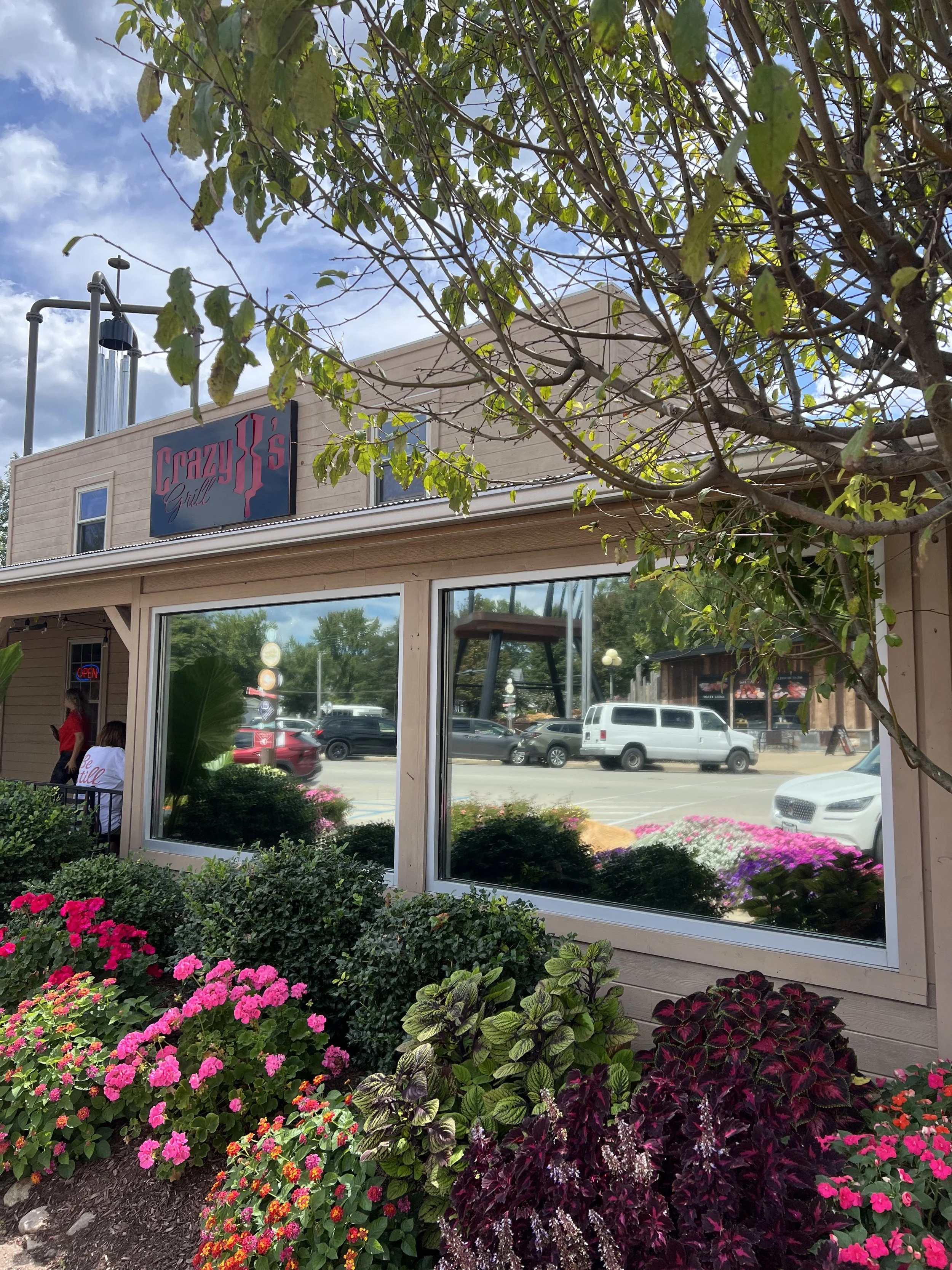 The exterior of Crazy J's Grill with a large window reflecting parked cars and buildings, surrounded by colorful flowers and greenery, with a sign on the building and a blue sky overhead.