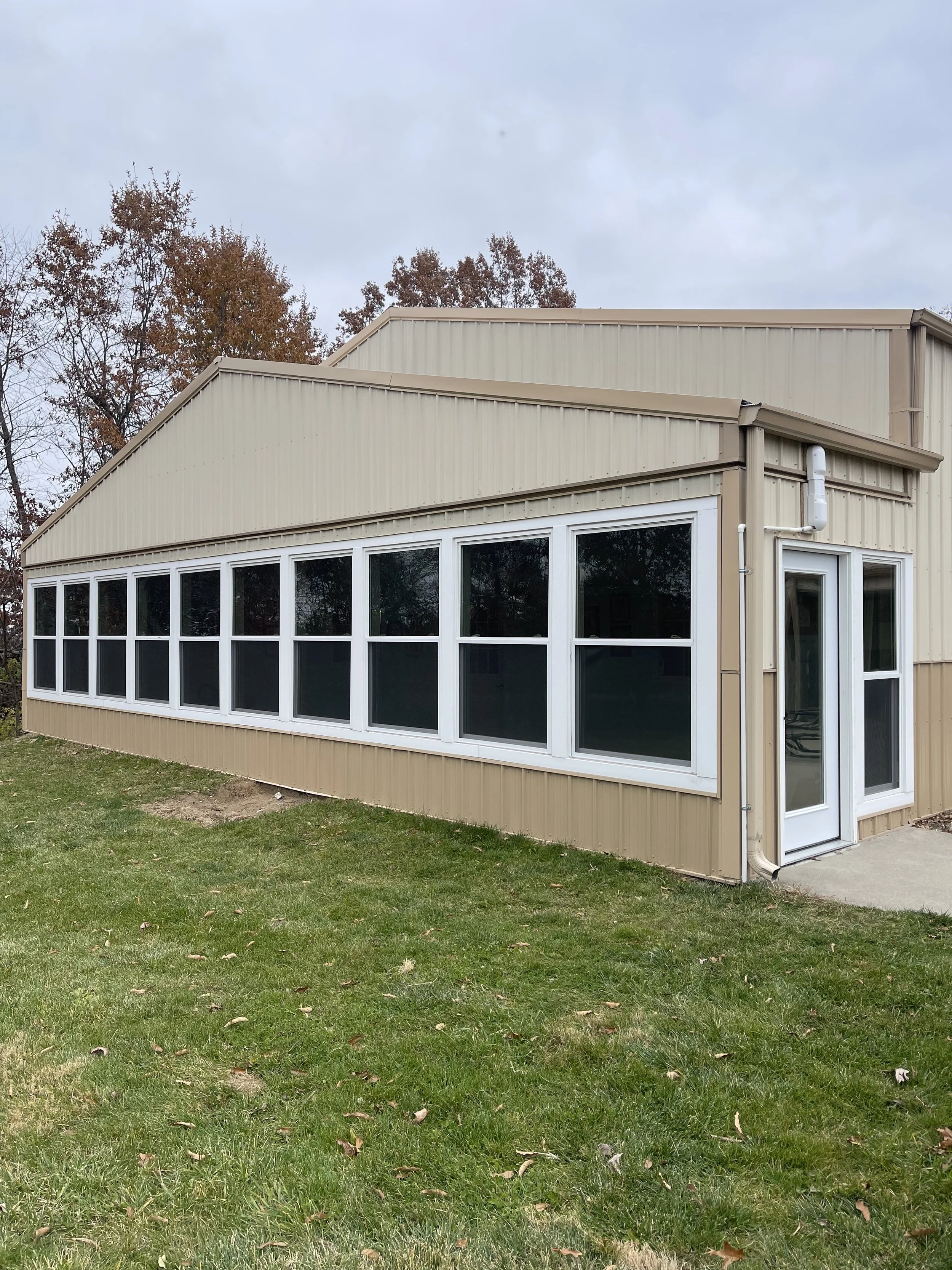 A beige metal building with a row of large windows and a door, set on a grassy area with trees and a cloudy sky in the background.