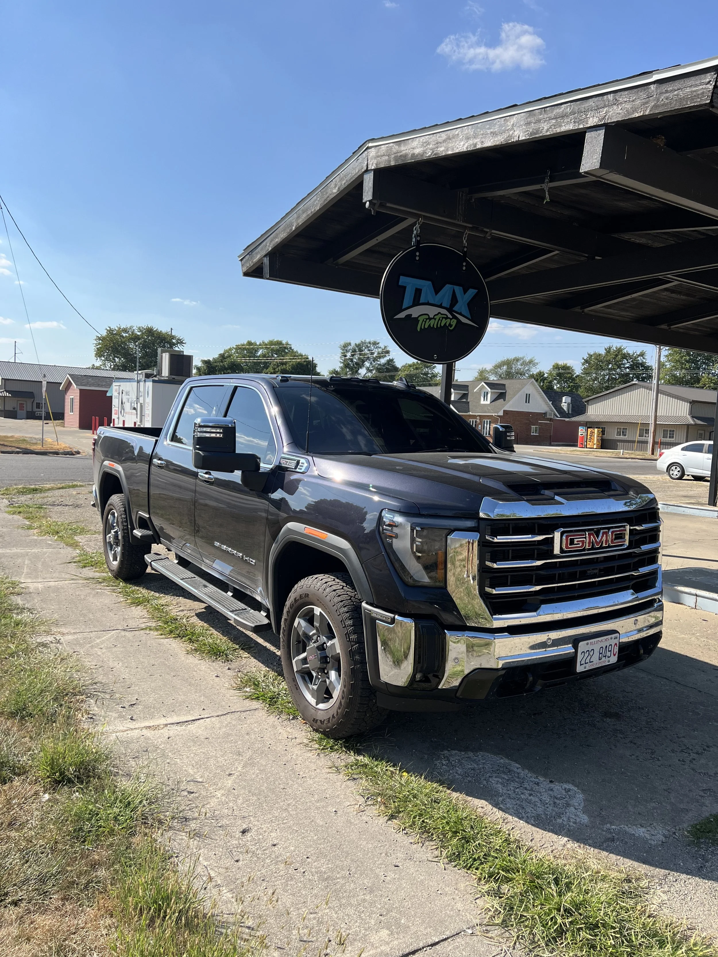 Black GMC Sierra HD pickup truck parked in front of a car wash or detailing shop with a sign that says "TMX Tinting" under a small roof extension.