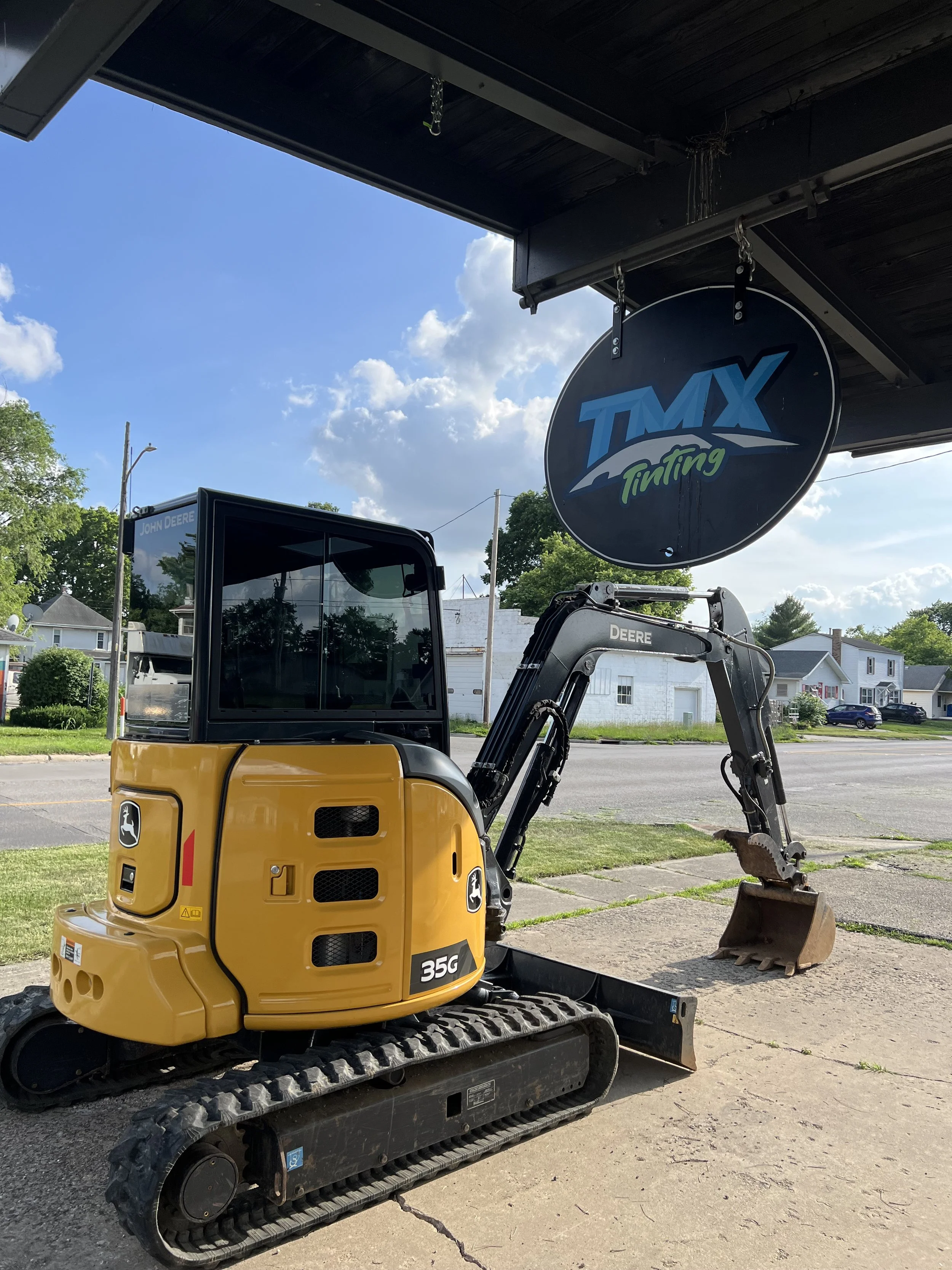 A yellow Deere 35G mini excavator parked outside under a roofing structure with a sign that says 'TMX Tuning' hanging from it. The background shows a street with houses and trees under a partly cloudy sky.